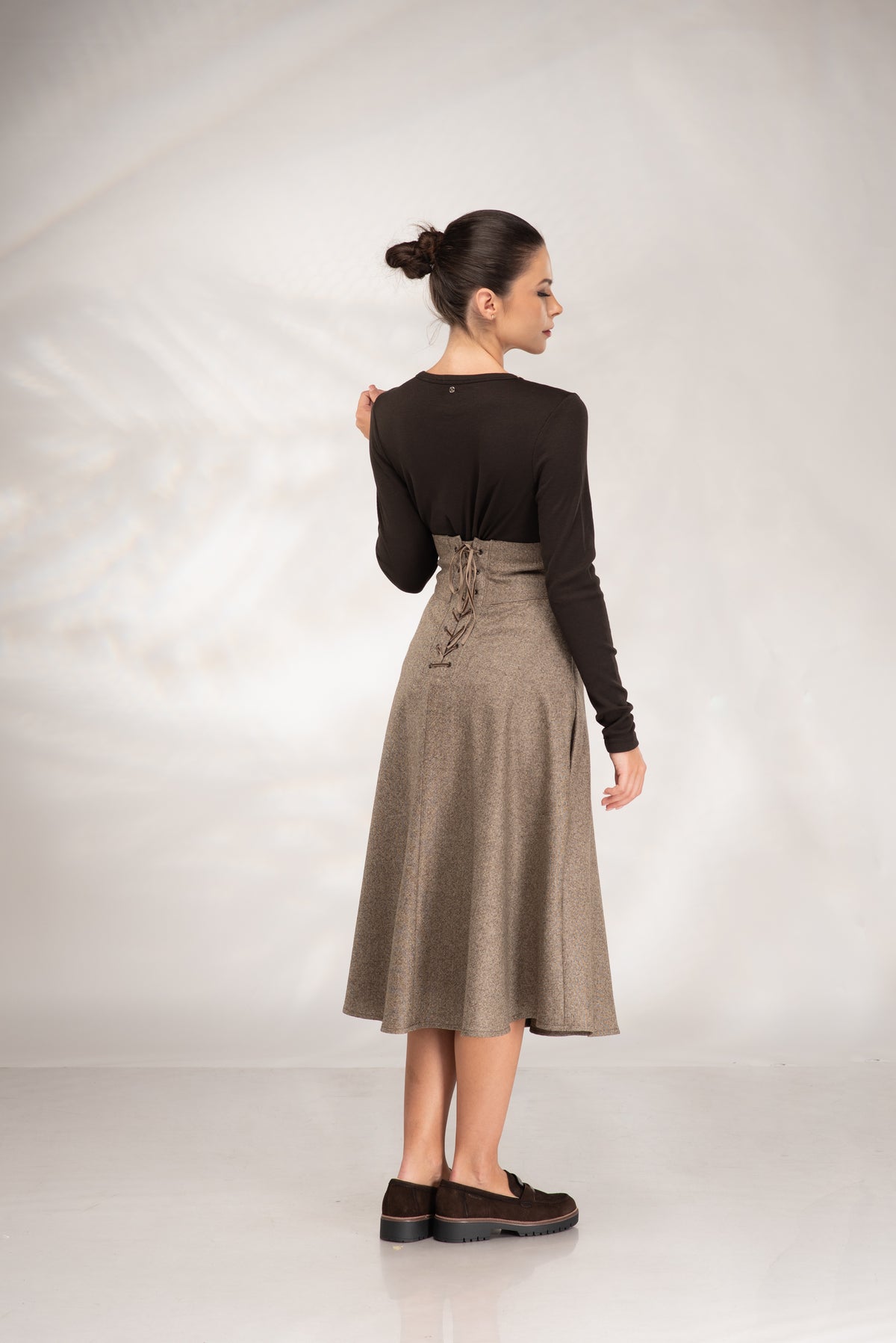 A woman is shown from the back wearing a black long-sleeve top and a high-waisted taupe-brown skirt with lace-up detailing at the back waist, paired with dark brown loafers, against a light gray studio backdrop.