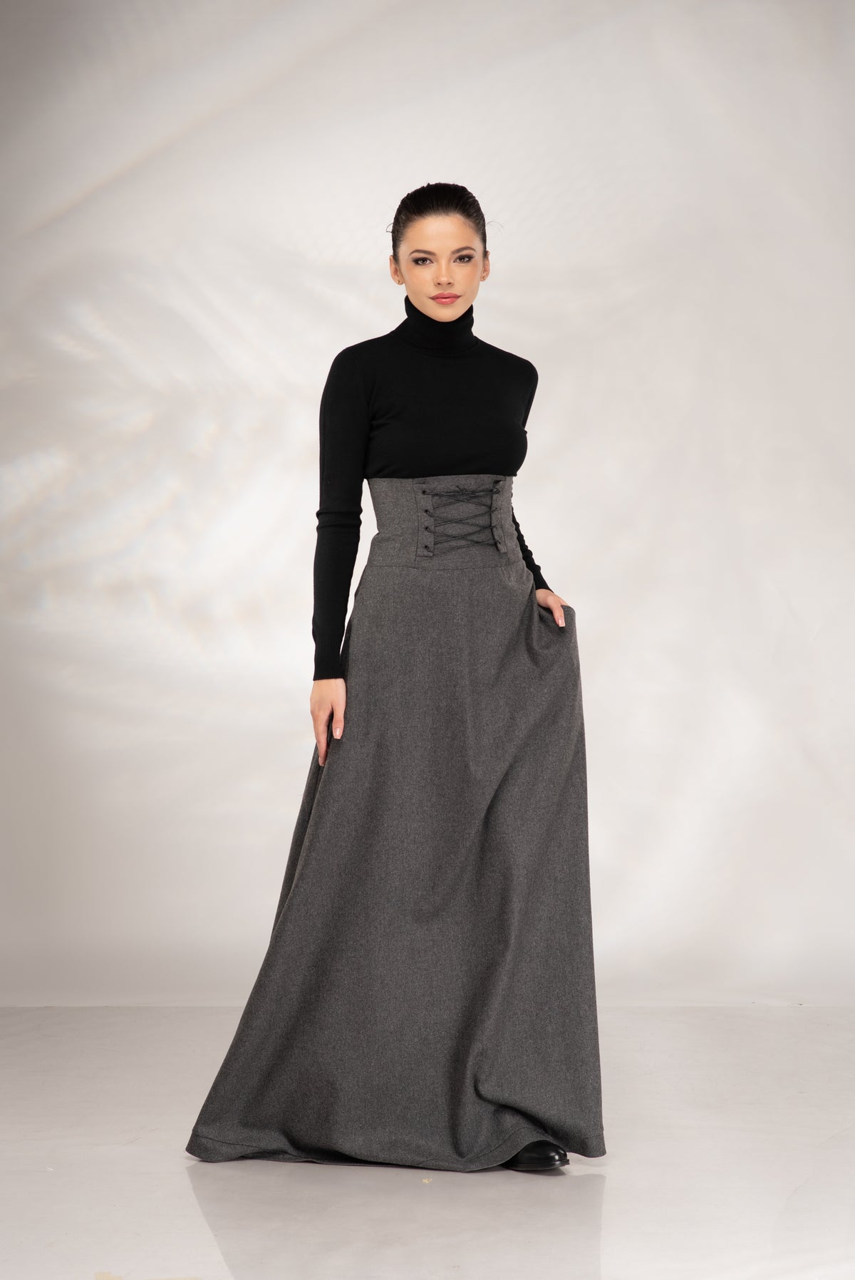 A model wearing a black long-sleeve mock neck top and a high-waisted, floor-length charcoal gray skirt with a lace-up corset-style waist, standing against a light gray studio backdrop.