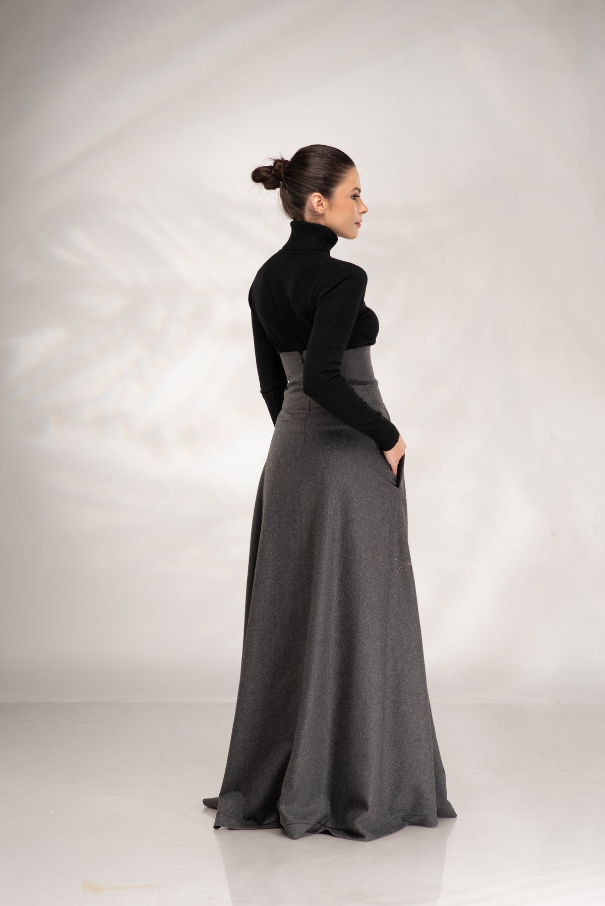 A woman wearing a long black mock-neck top and a high-waisted, floor-length dark gray skirt with side pockets, shown from a three-quarters back view against a light gray studio backdrop.