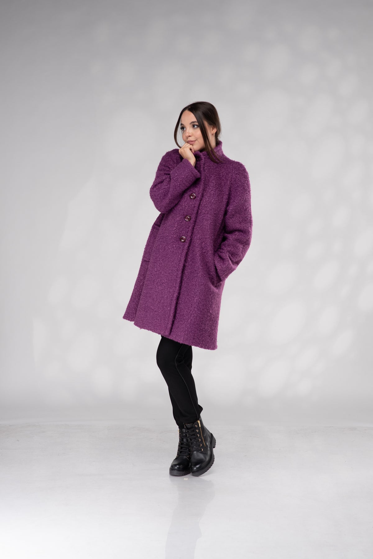 A woman modeling a textured, fuzzy purple coat with black pants and black boots, standing against a light gray studio background with soft shadow patterns.