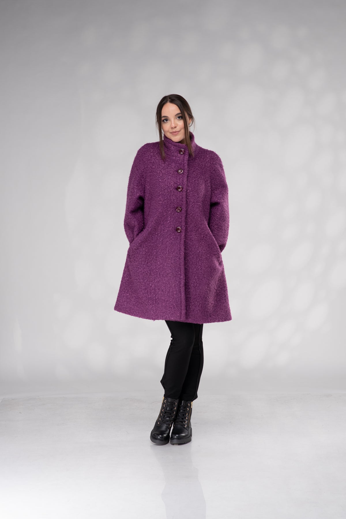 A woman modeling a textured purple wool-blend coat with dark buttons, paired with black pants and black lace-up ankle boots, standing against a light gray studio backdrop.
