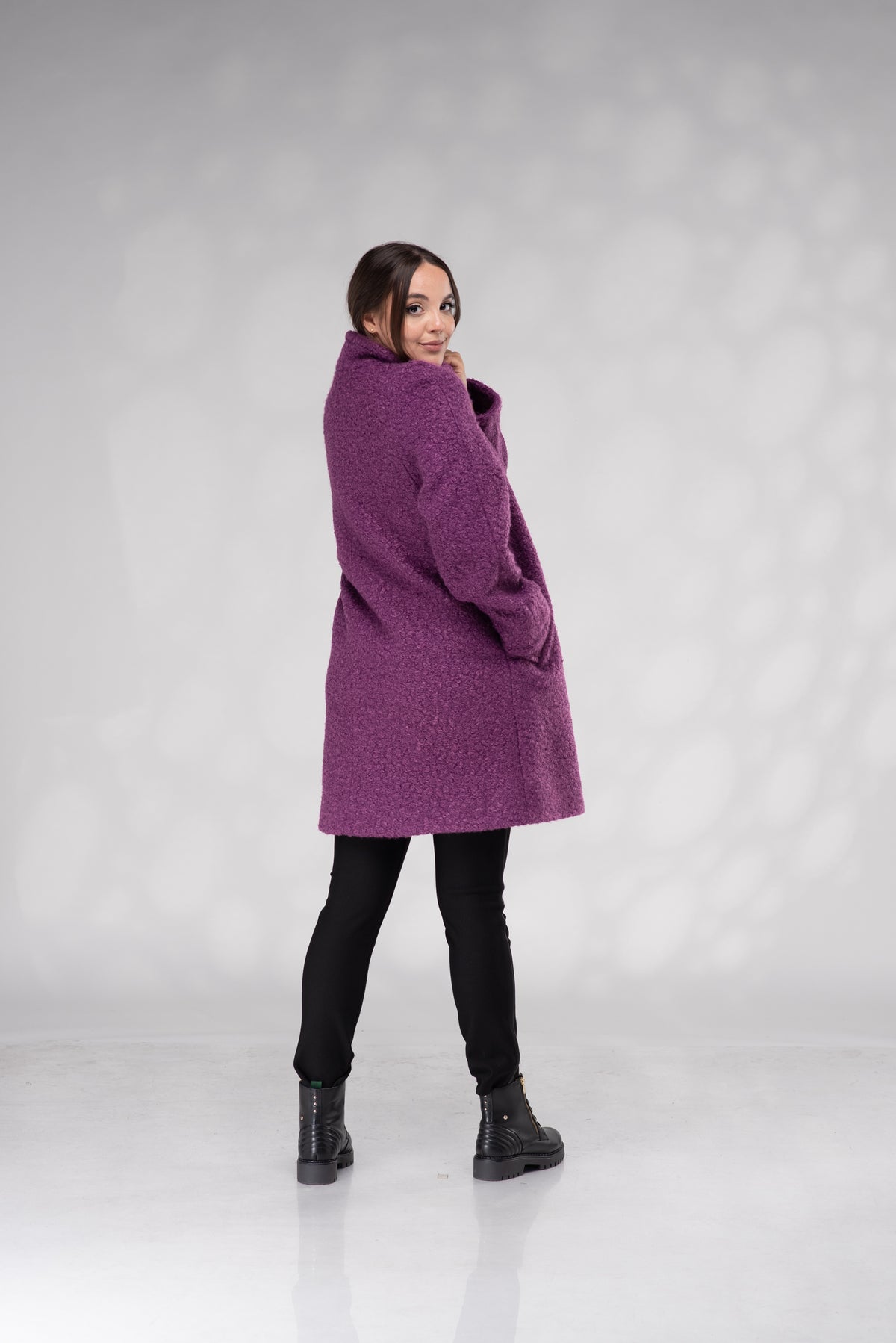 A woman modeling a textured, fuzzy purple mid-length coat with a high collar, paired with black pants and black boots, standing against a light gray studio backdrop.