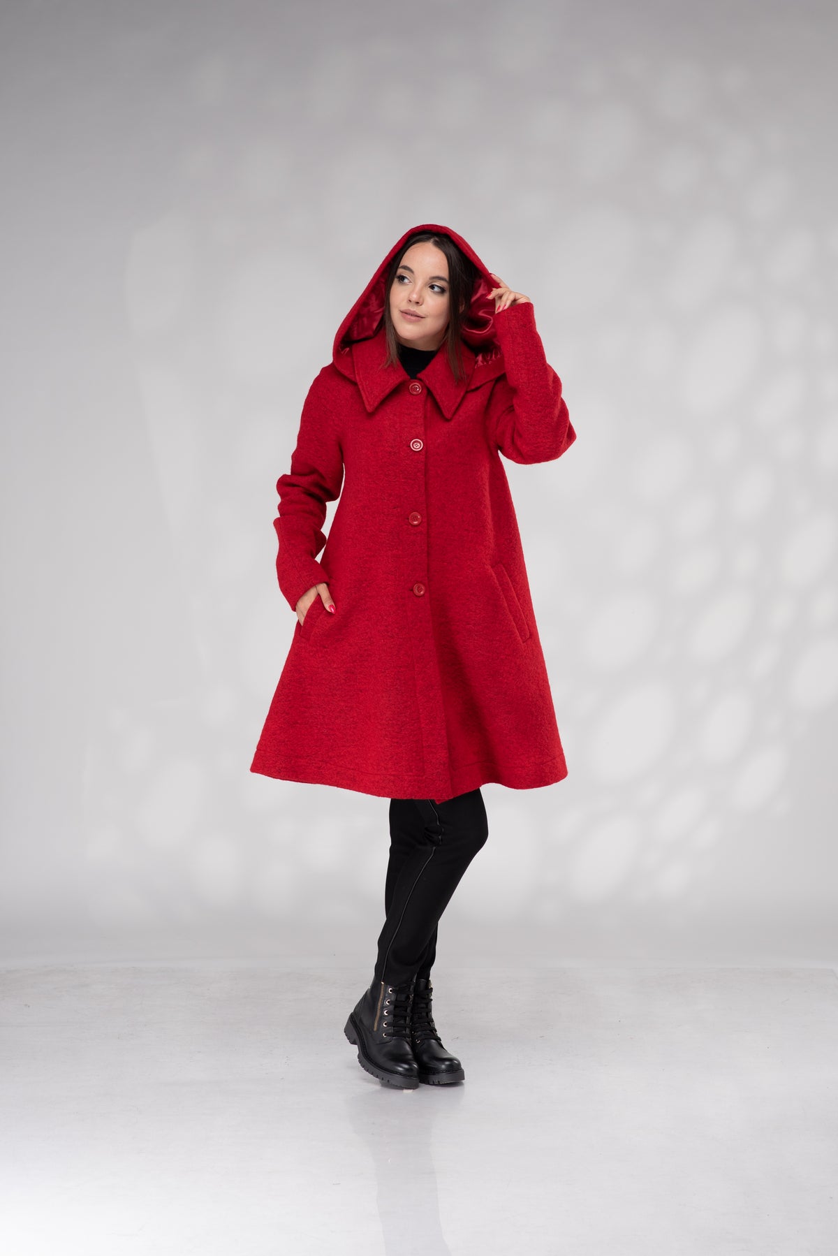 A woman modeling a bright red hooded coat with a wide collar and front buttons, paired with black tights and black boots, standing against a light gray studio backdrop with soft circular shadows.
