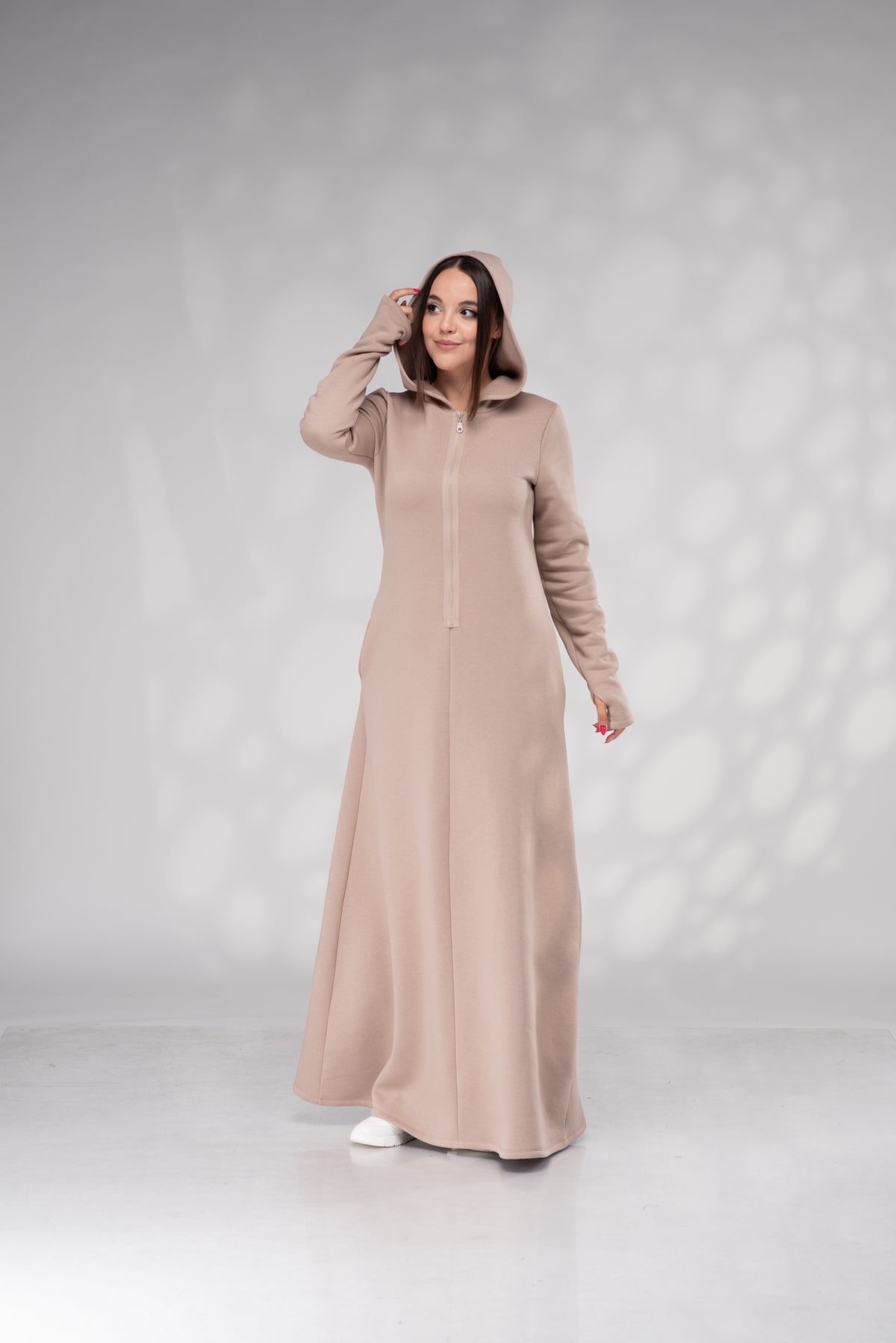 A woman modeling a long, hooded maxi dress in a solid light taupe-beige color, standing against a light gray studio backdrop with soft shadow patterns.
