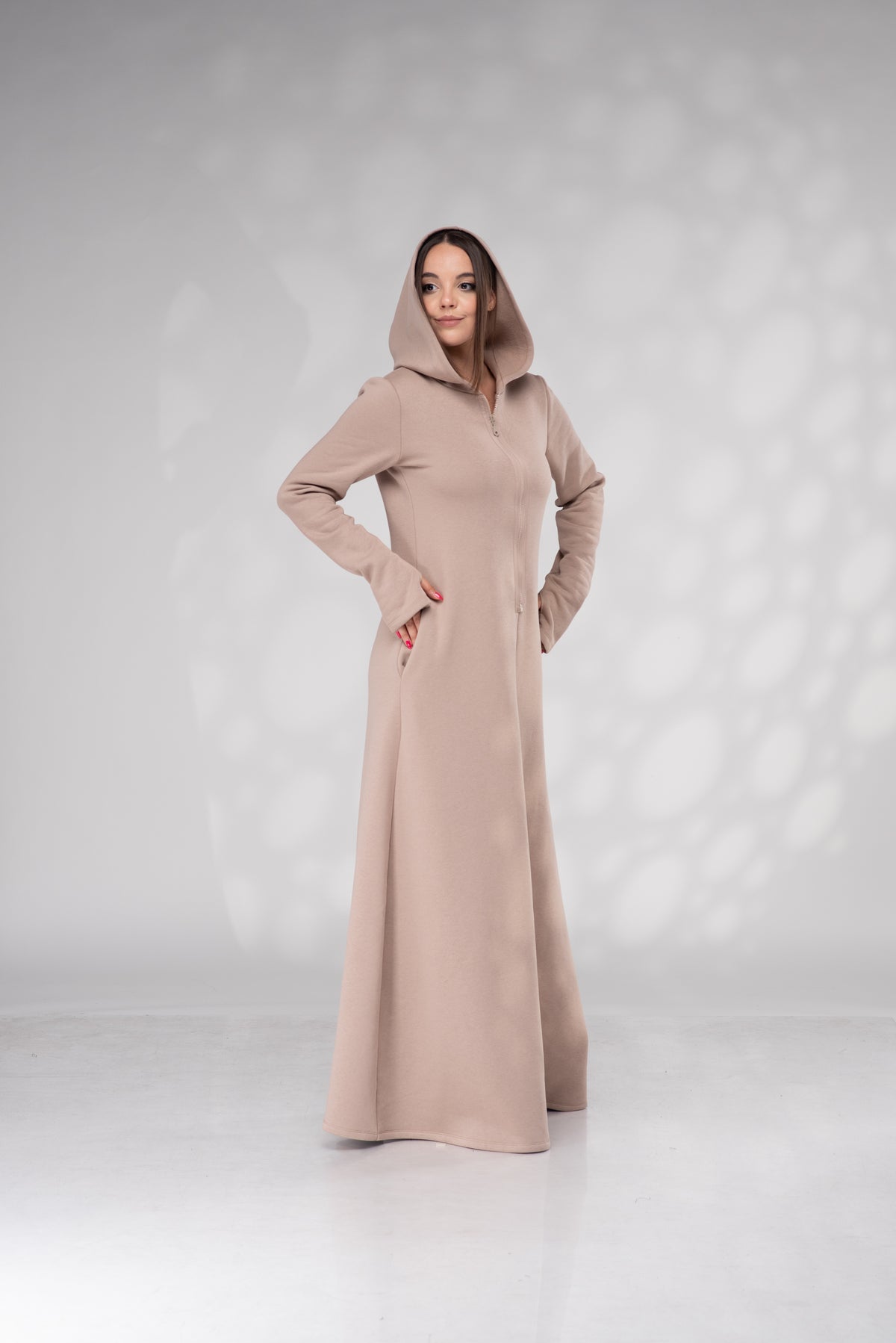 A woman modeling a long, floor-length hooded dress in a light taupe-beige color, standing against a light gray studio backdrop with soft shadow patterns.