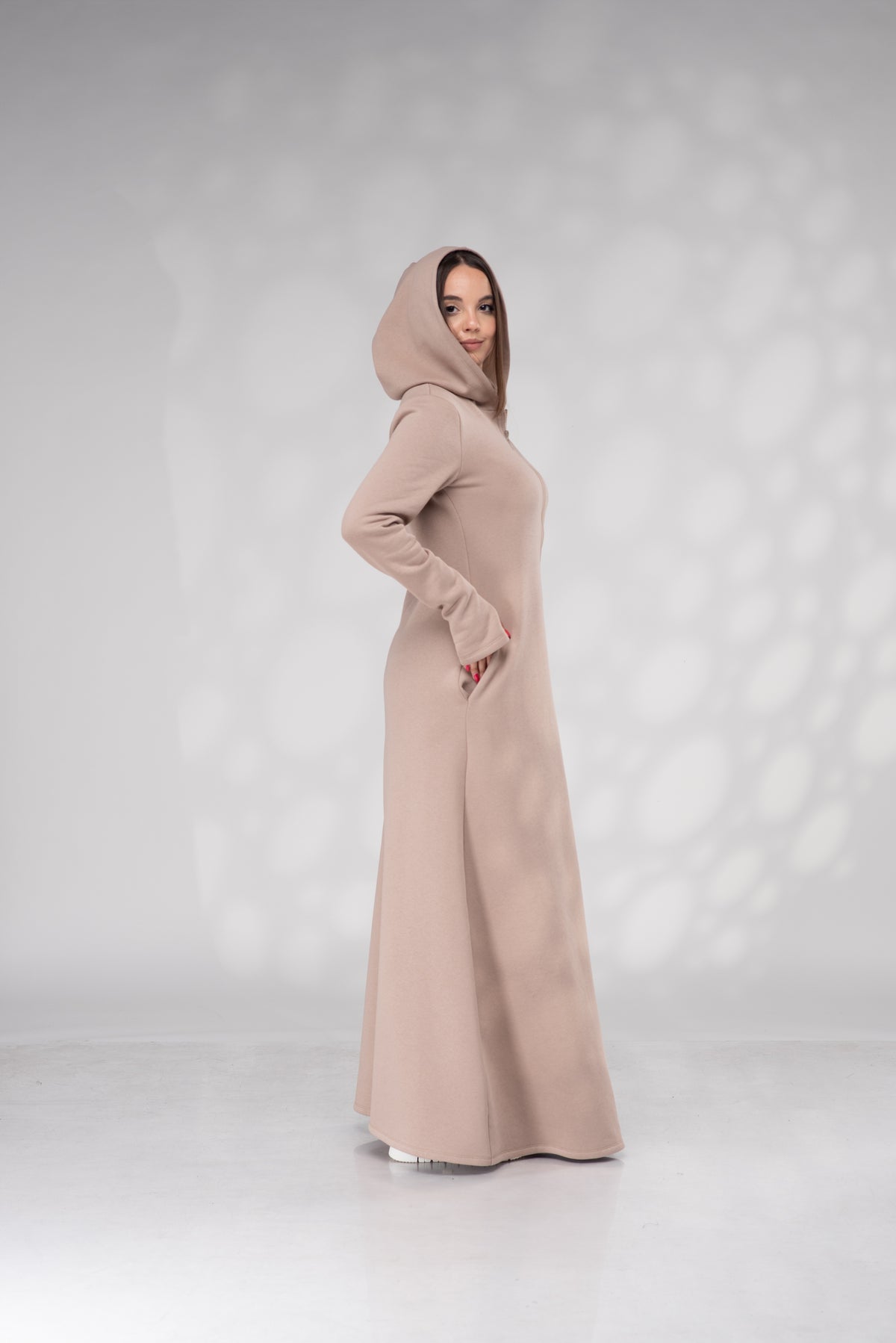 A woman wearing a long, floor-length beige hooded abaya with long sleeves and side pockets, standing in profile against a light gray studio backdrop with soft shadow patterns.