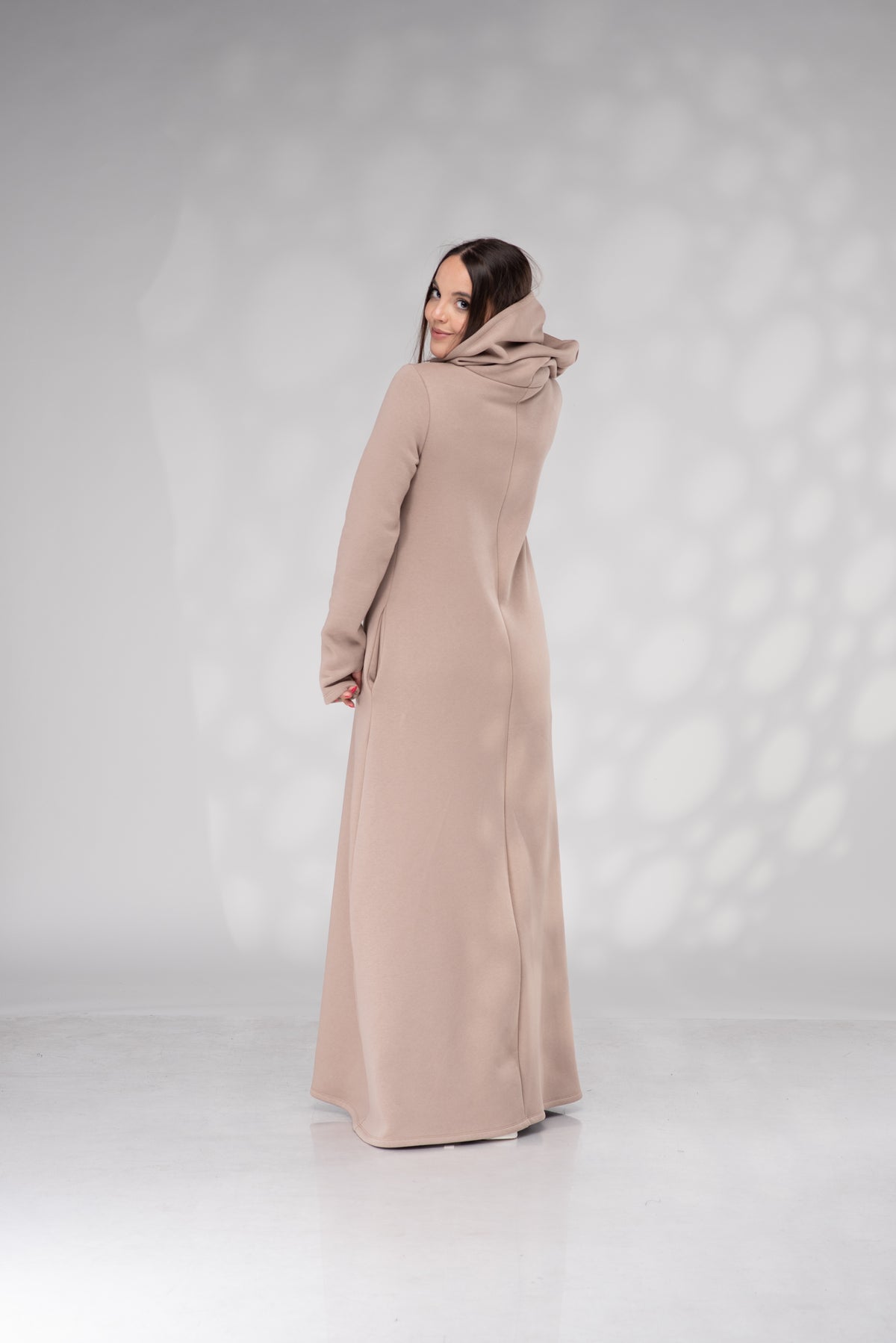 A woman wearing a long, floor-length, draped hooded coat in a light taupe-beige color, photographed from the back against a light gray studio backdrop with soft shadow patterns.