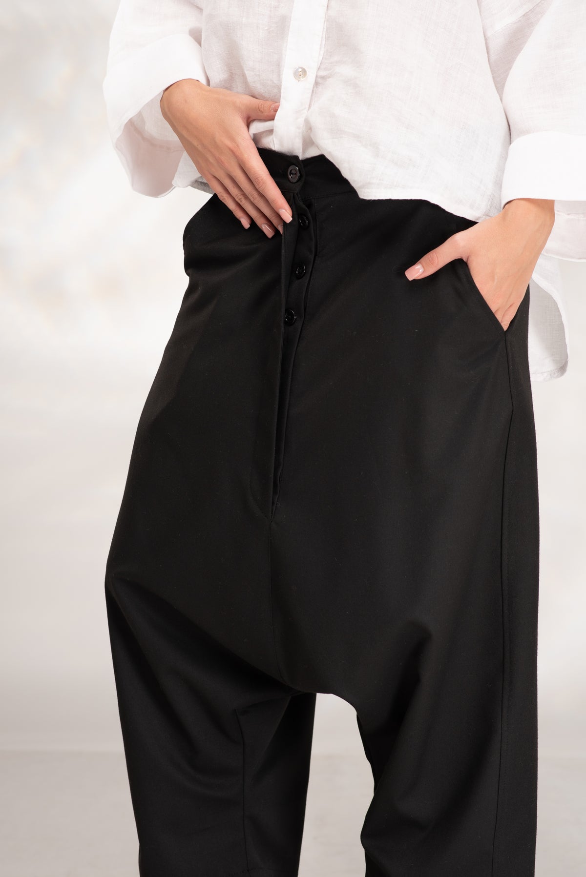 A close-up of a person wearing black baggy pants with a front button closure and side pockets, paired with a white button-up shirt. The person's hands are tucked into the pockets.