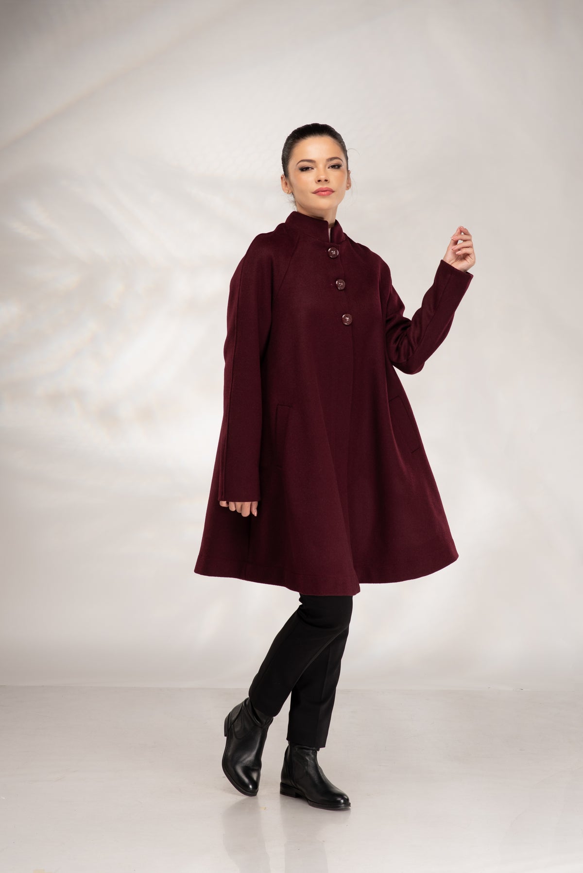 A woman modeling a long-sleeved, high-neck, knee-length coat in deep burgundy. The coat features a buttoned front and a flared silhouette, paired with black pants and black ankle boots, set against a light gray studio backdrop.