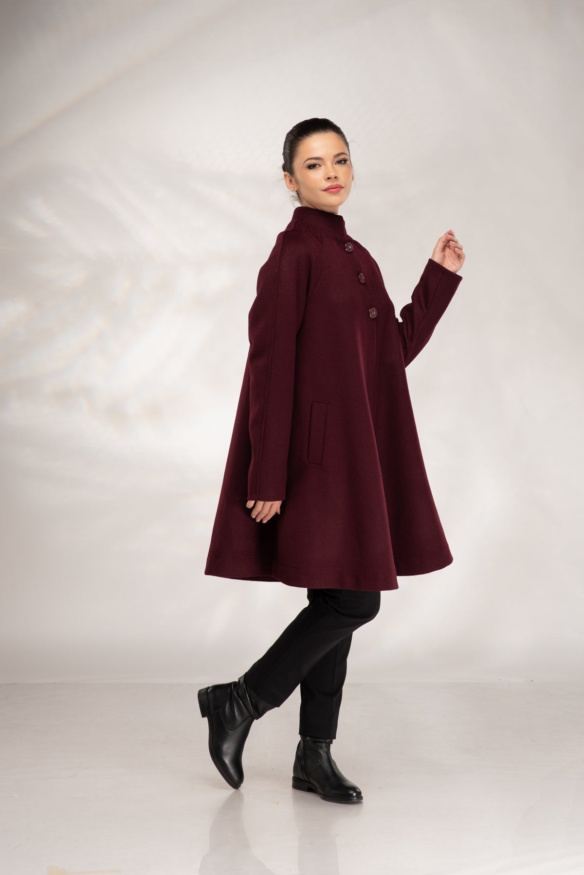 A woman modeling a long, high-neck, knee-length coat in deep burgundy. The coat features a structured silhouette, long sleeves, and a buttoned front.