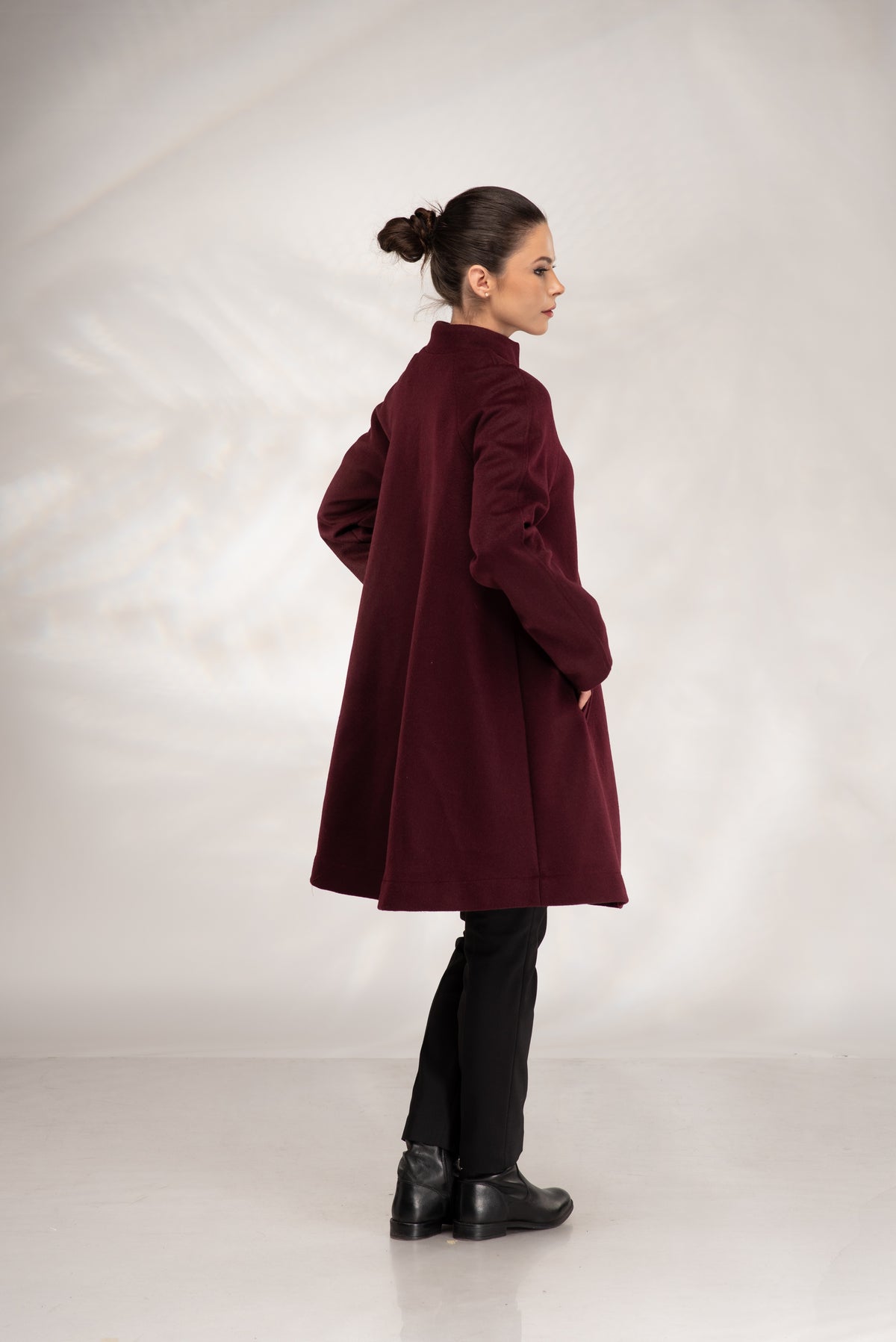 A woman wearing a long, dark burgundy coat, black pants, and black boots, standing in profile against a light gray studio backdrop.