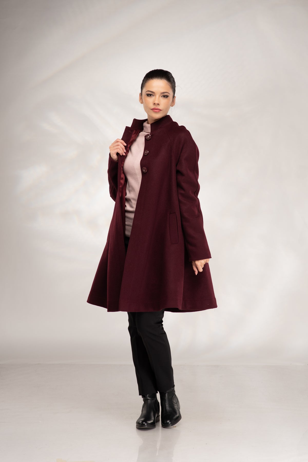 A woman modeling a long, dark burgundy wool coat with a high collar, worn over a light pink top, black pants, and black boots, standing against a light gray studio backdrop.