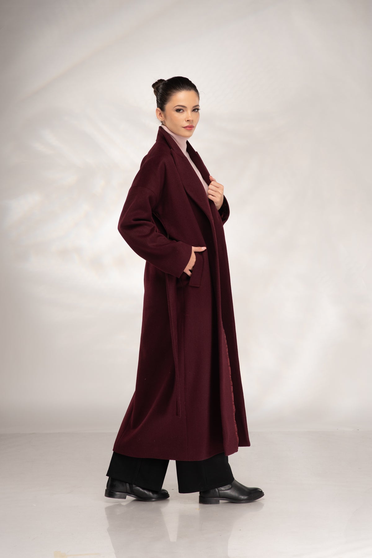 A woman modeling a long, open-front burgundy coat over a light pink top and black pants, standing against a light gray studio backdrop.