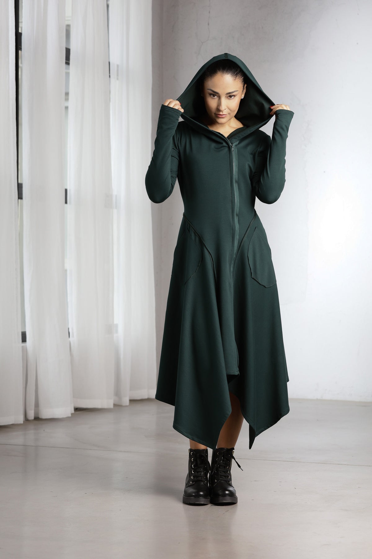 A woman wearing a dark forest green hooded zip-up dress with asymmetrical hem and front pockets, paired with black lace-up ankle boots, standing in a bright studio with white curtains.