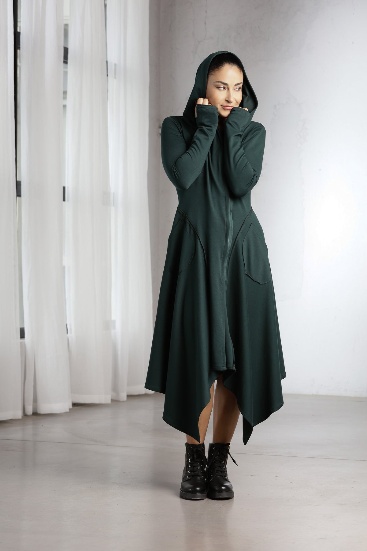 A woman wearing a dark forest green hooded asymmetrical long coat with black combat boots, standing in a minimalist studio with white curtains and a light gray wall.