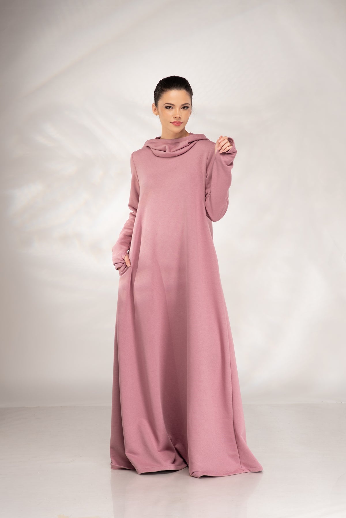 A woman modeling a long-sleeved, floor-length dusty pink dress with a draped cowl neckline, standing against a light gray studio backdrop.