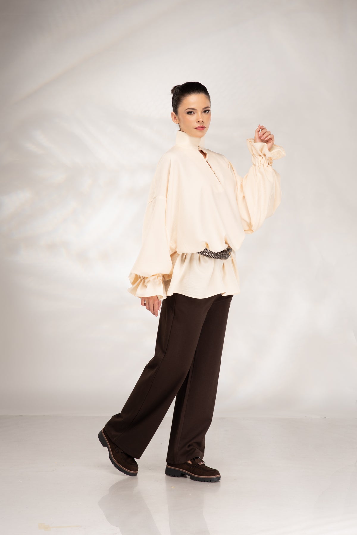 A model posing in a studio setting wearing a cream-colored, long-sleeved blouse with dramatic ruffled sleeves and dark brown wide-leg trousers.