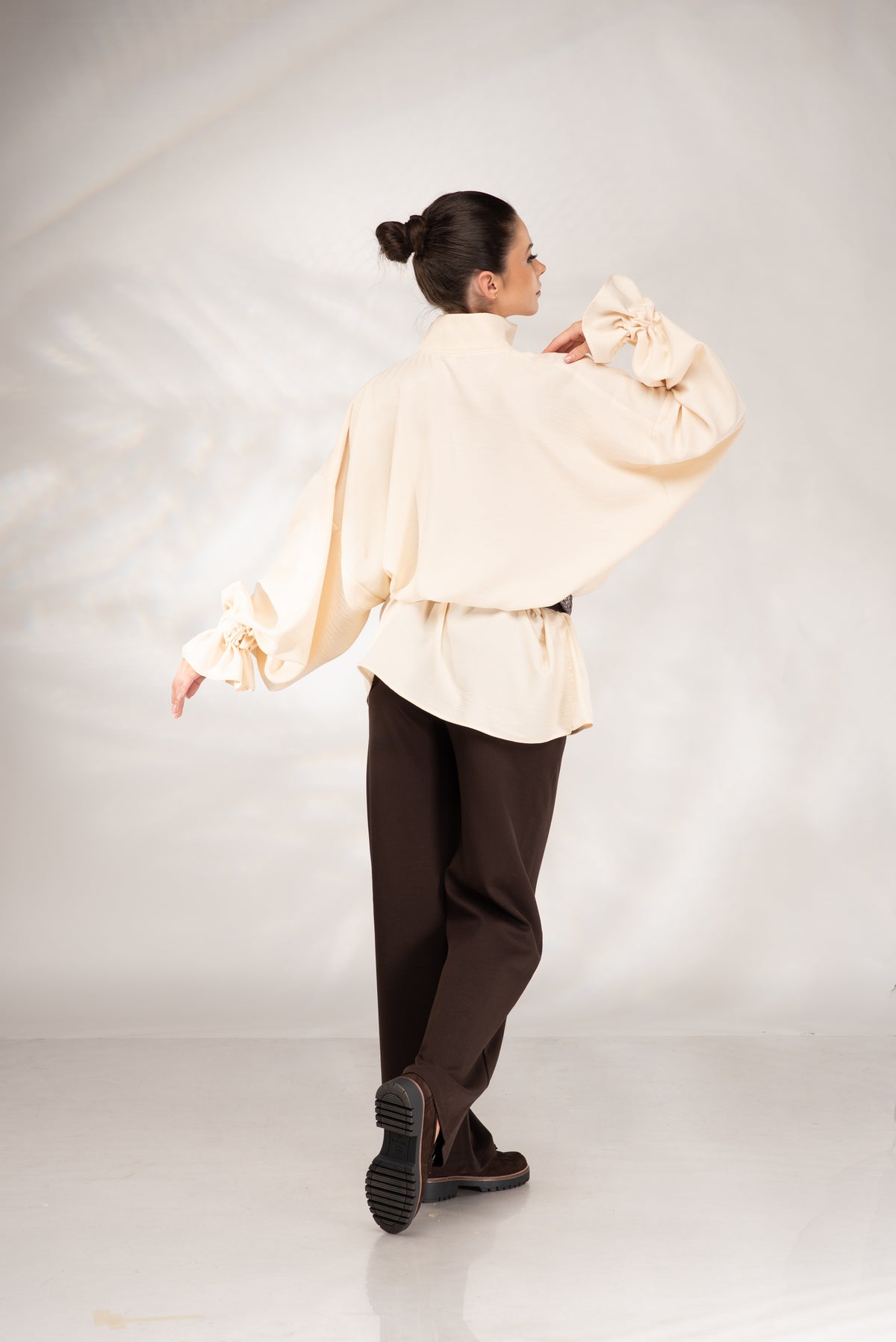 A woman is shown from behind wearing a cream-colored, oversized long-sleeve blouse with dramatic gathered sleeves and dark brown trousers, standing against a light gray studio backdrop.