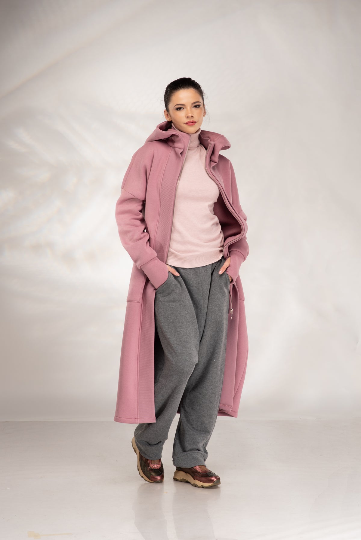 A woman modeling a long, dusty pink hooded coat over a light pink turtleneck sweater, gray sweatpants, and brown shoes, standing against a light gray studio backdrop.