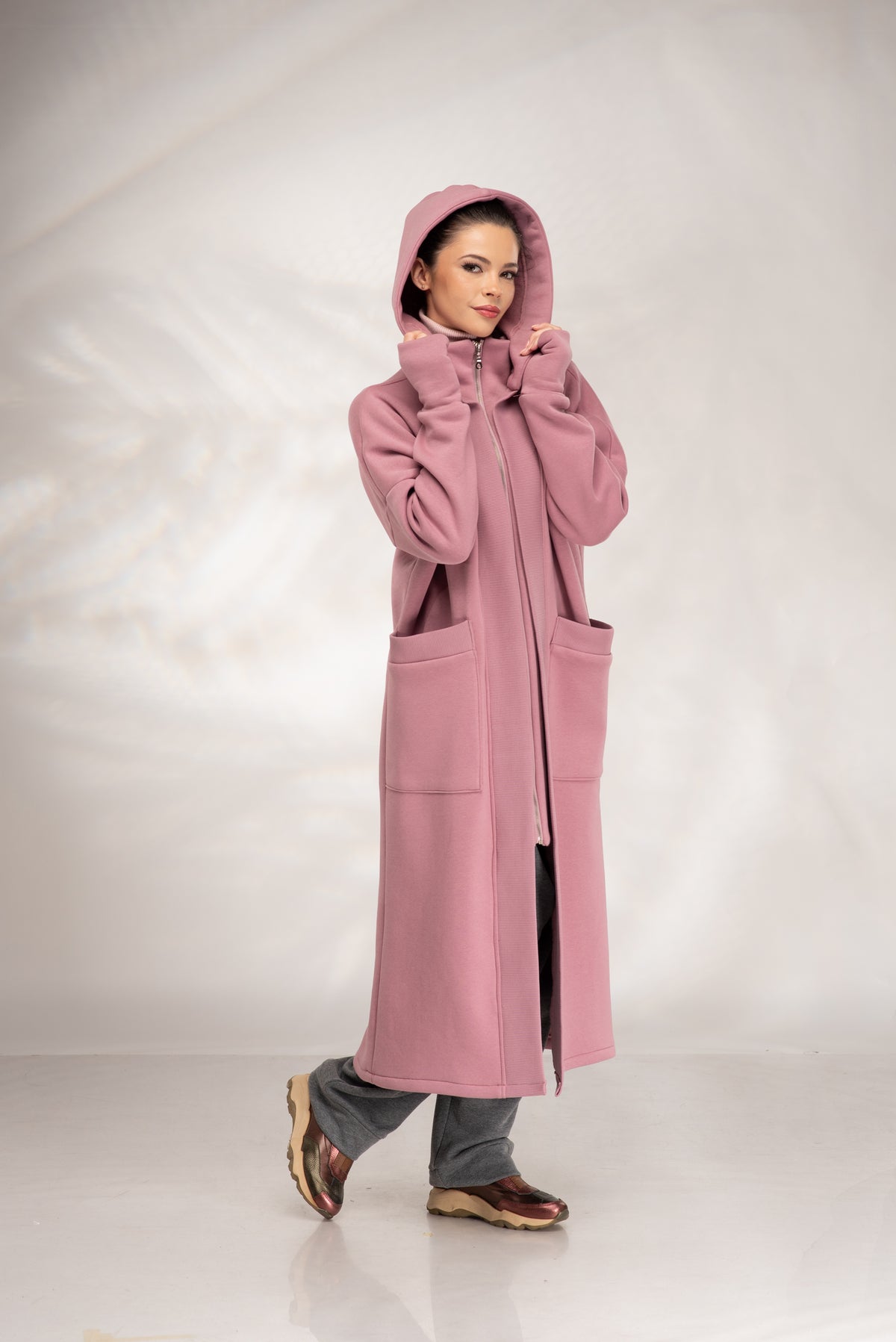 A woman wearing a long, hooded pink coat with large front pockets, styled with gray pants and brown shoes, standing against a light gray studio backdrop.