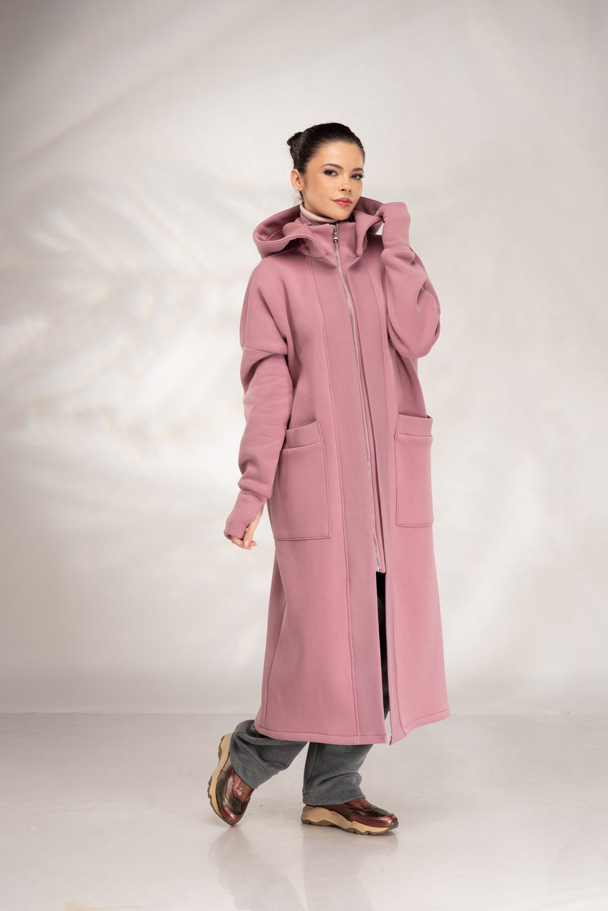 A woman modeling a long, dusty pink hooded coat with a front zipper and large patch pockets, paired with gray pants and brown shoes, against a light gray studio backdrop.