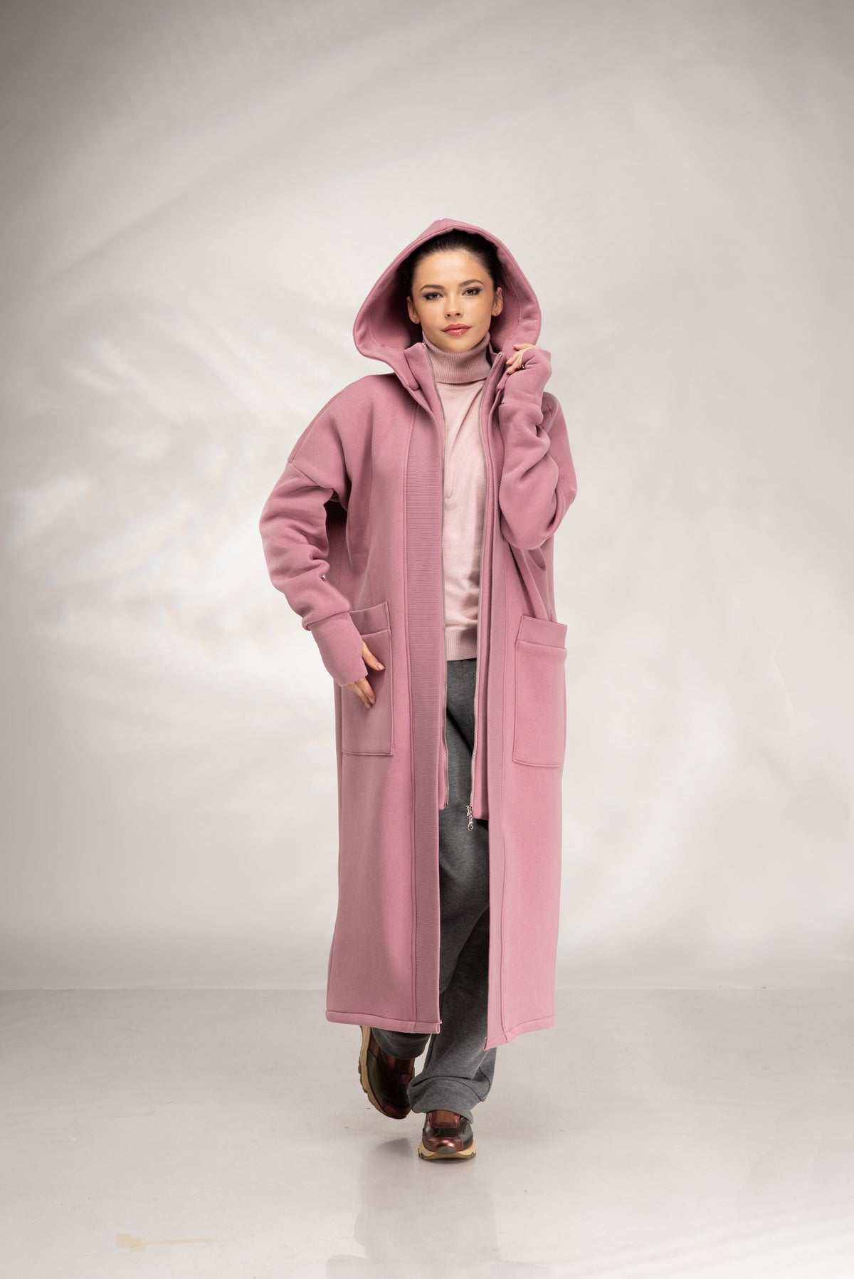 A model wearing a long, dusty pink hooded coat over a light pink turtleneck sweater, gray pants, and brown shoes, standing against a light gray studio backdrop.