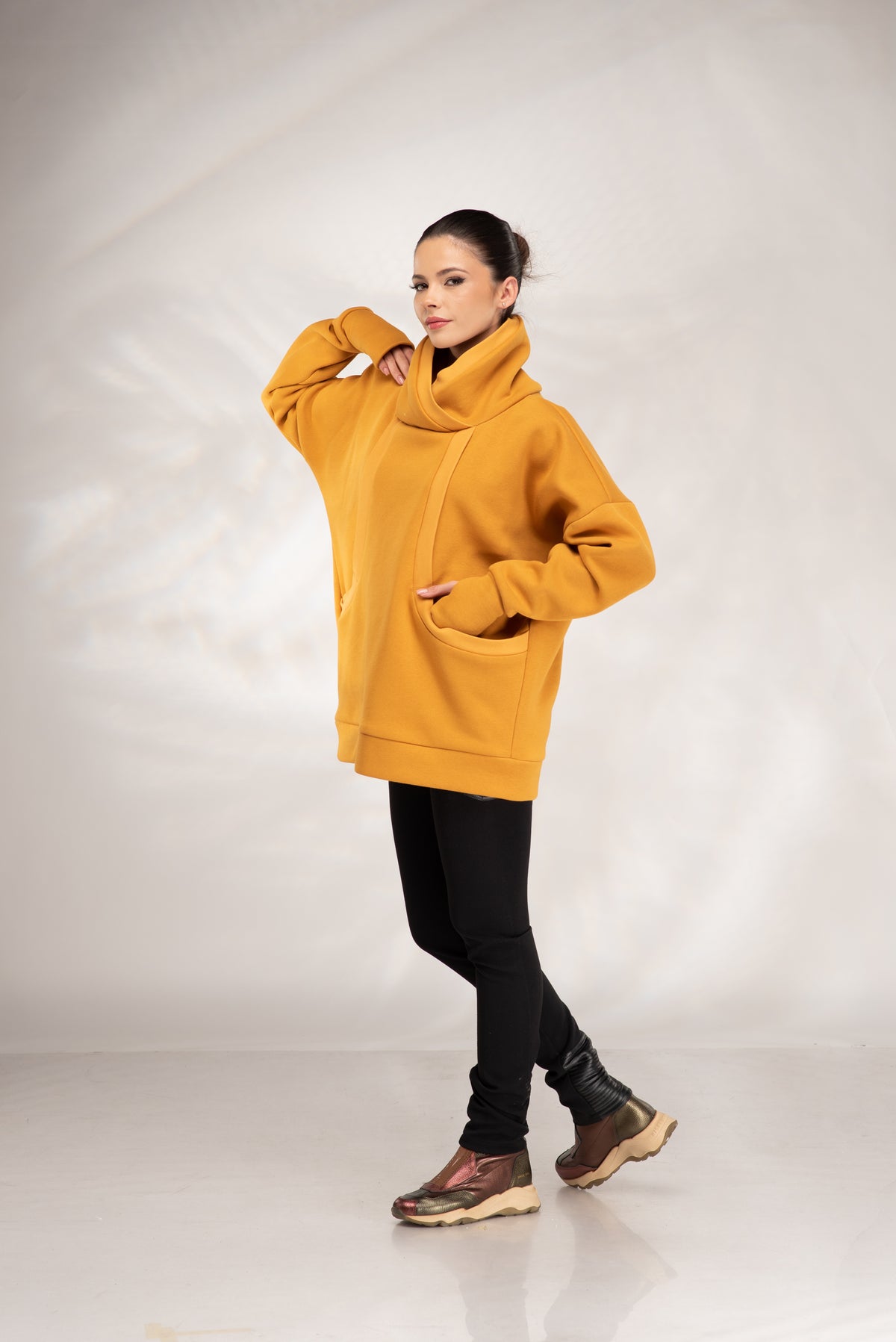 A woman modeling an oversized mustard yellow cowl-neck hoodie with front pockets, paired with black pants and brown hiking boots, against a light gray studio backdrop.