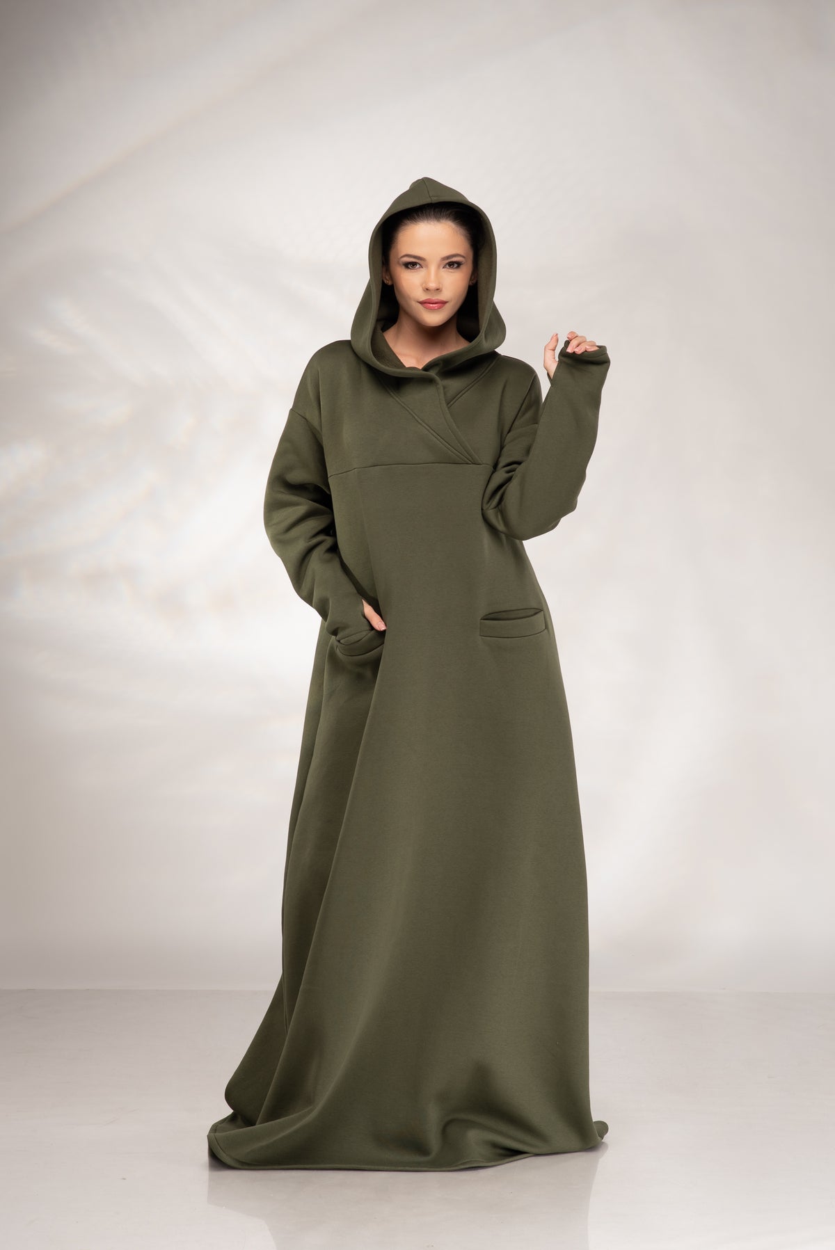 A woman modeling a long, loose hooded robe in olive green, standing against a light gray studio backdrop.