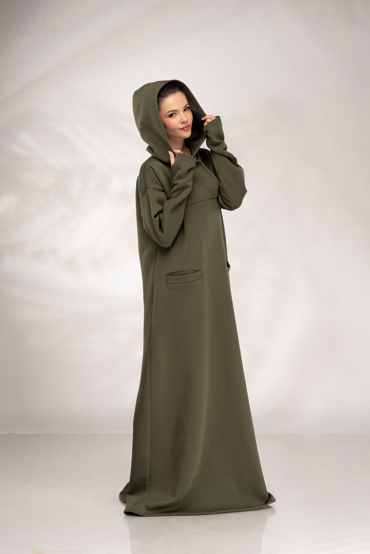 A woman modeling a long, flowing olive green hooded robe or dress with a front pocket, standing against a light gray studio backdrop.