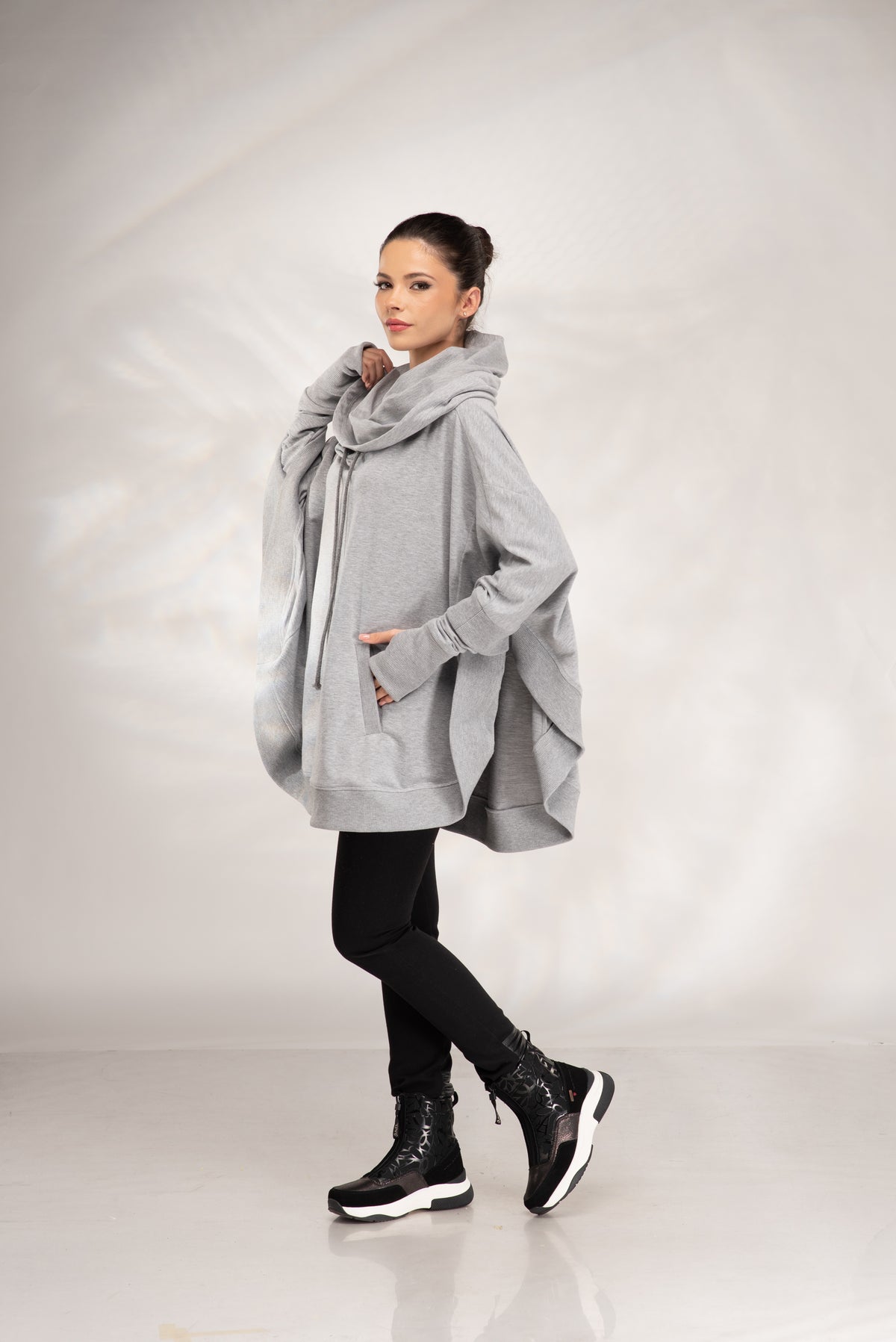 A woman modeling a light gray oversized hooded sweatshirt with a draped, layered design, paired with black pants and black sneakers, against a light gray studio background.
