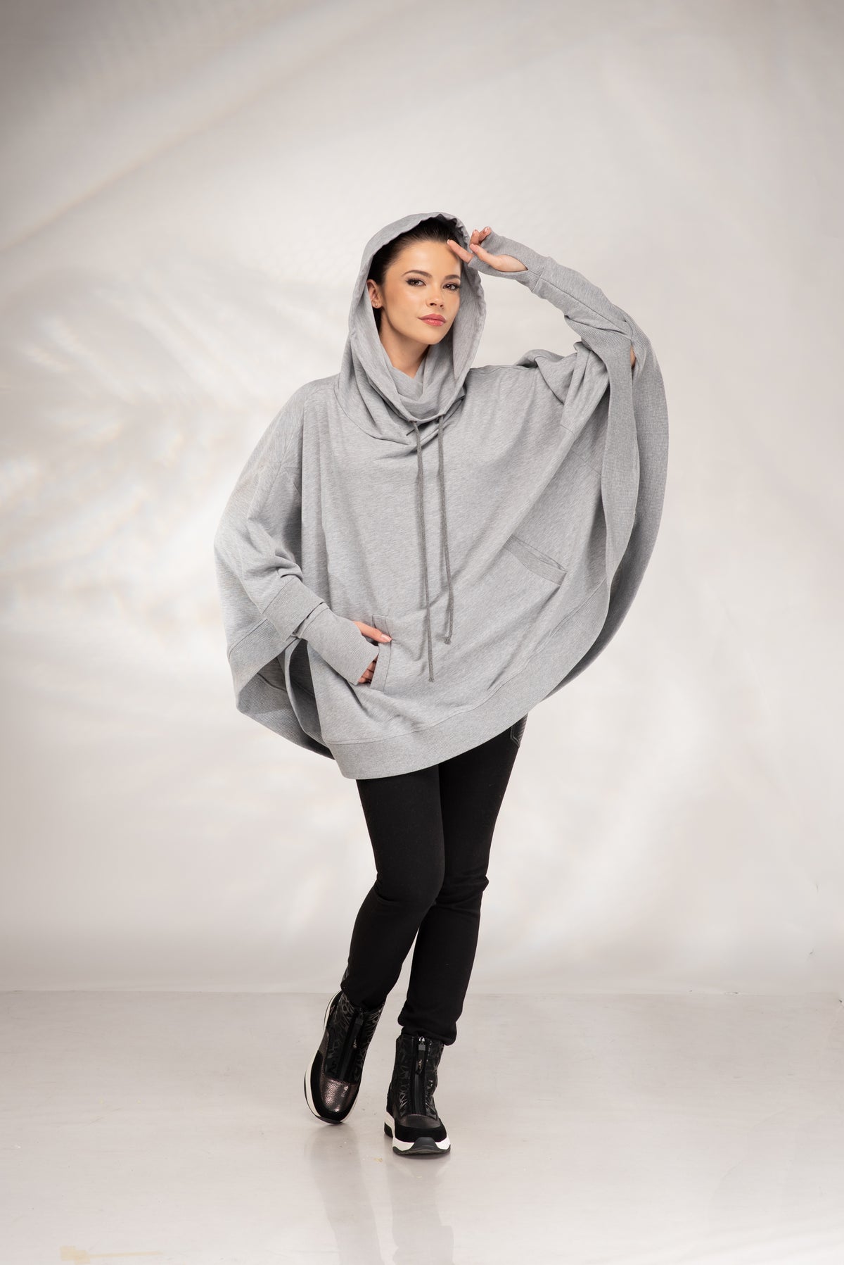 A woman modeling a light gray oversized poncho hoodie with a high draped collar and drawstrings, styled with black pants and black boots against a light gray studio backdrop.