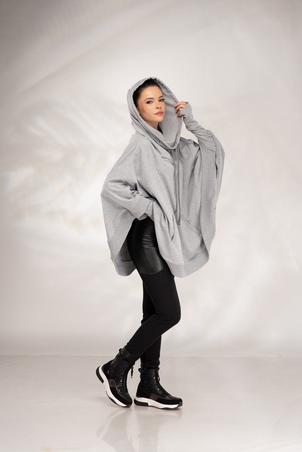 A woman modeling an oversized light gray hooded poncho-style jacket, paired with black leggings and black boots, against a light gray studio backdrop.