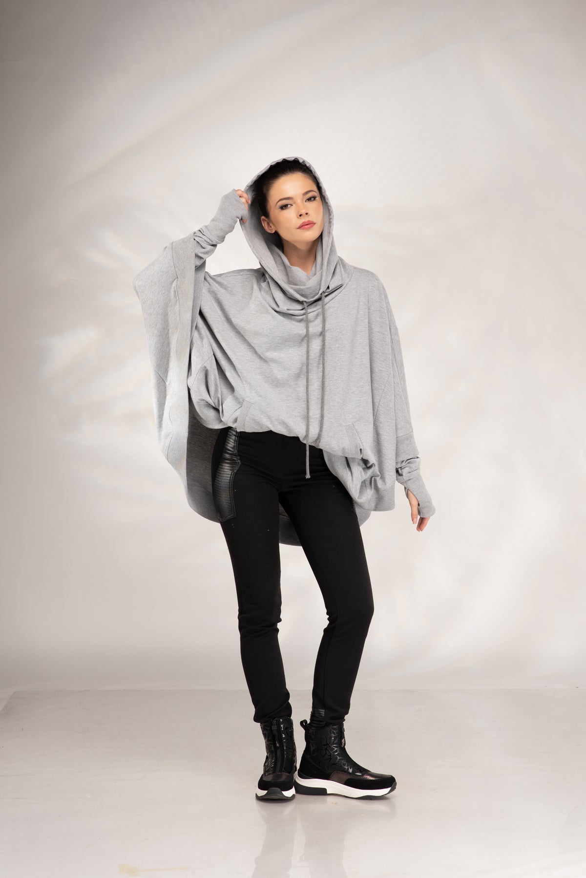 A model wearing a light gray oversized draped hoodie, black skinny pants, and black lace-up boots, standing against a light gray studio backdrop.