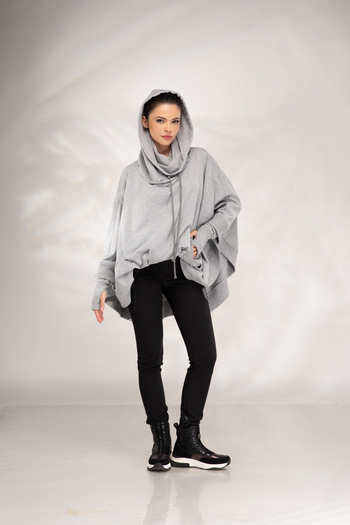 A woman modeling a light gray oversized poncho-style top with a draped hood, paired with black pants and black lace-up boots, against a light gray studio backdrop.