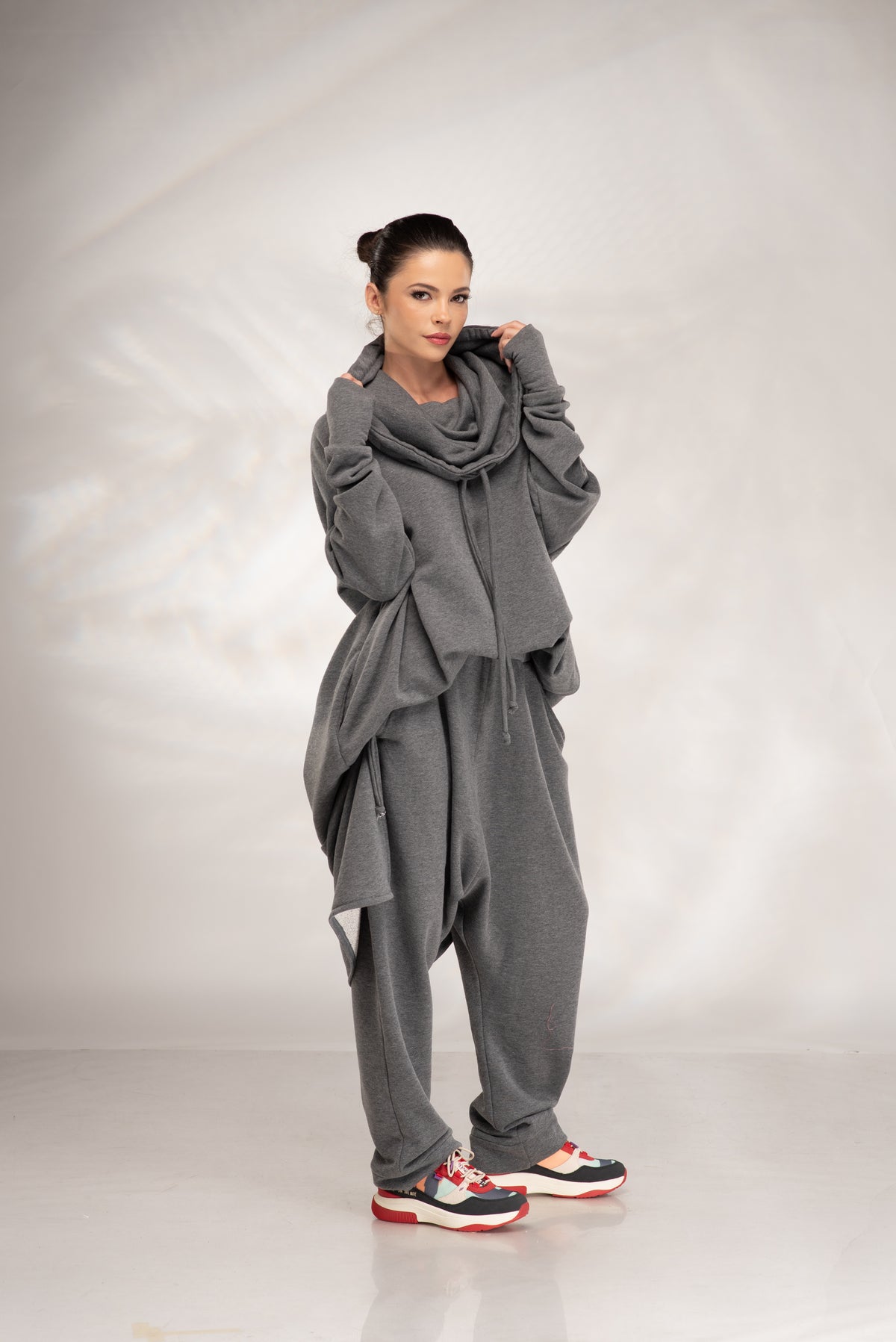 A woman modeling a gray lounge set consisting of a draped hoodie, a thick cowl neck scarf, and matching sweatpants, paired with colorful sneakers, against a light gray studio backdrop.