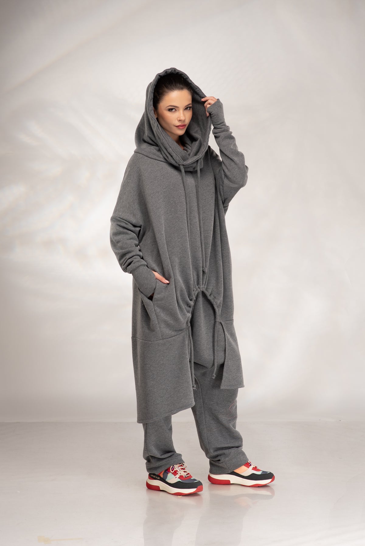 A woman modeling a long, oversized gray hooded sweatshirt with a matching pair of loose gray pants and colorful sneakers, standing against a light gray studio backdrop.