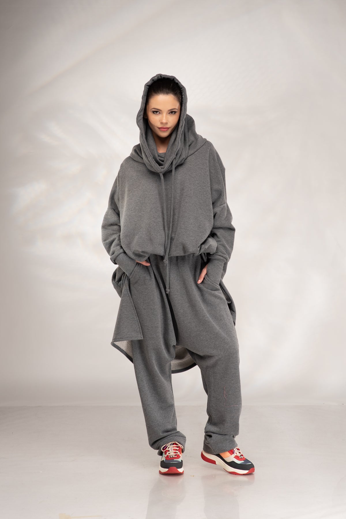A model wearing a loose-fitting, oversized gray hooded sweatshirt and matching sweatpants, styled with red and black sneakers, standing against a light gray studio backdrop.