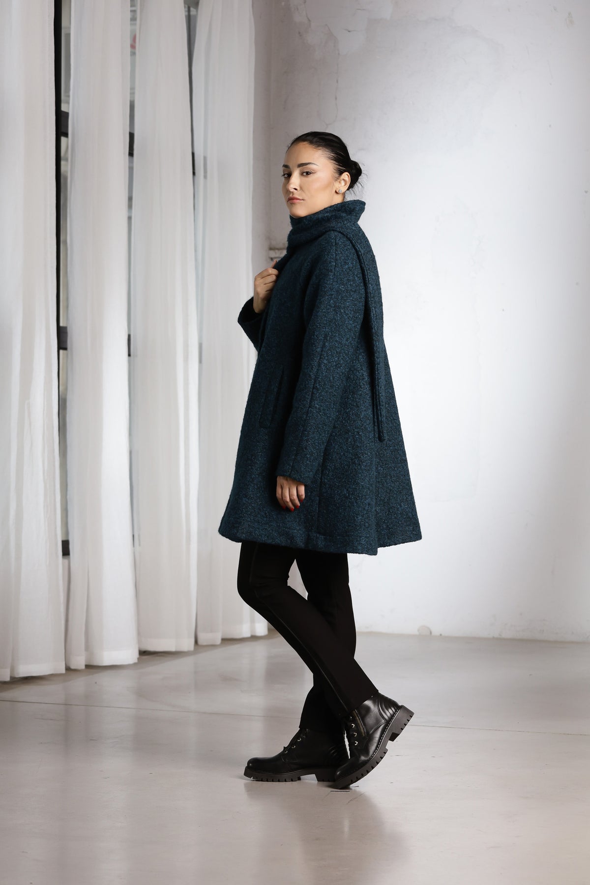 A woman wearing a dark teal textured coat, black tights, and black boots, standing in a minimalist studio with white curtains and a white wall.