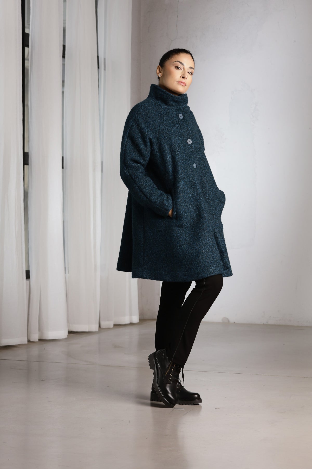 A woman modeling a dark teal blue textured coat with a high mock neck and front buttons, paired with black pants and boots, standing in a minimalist studio with white curtains.