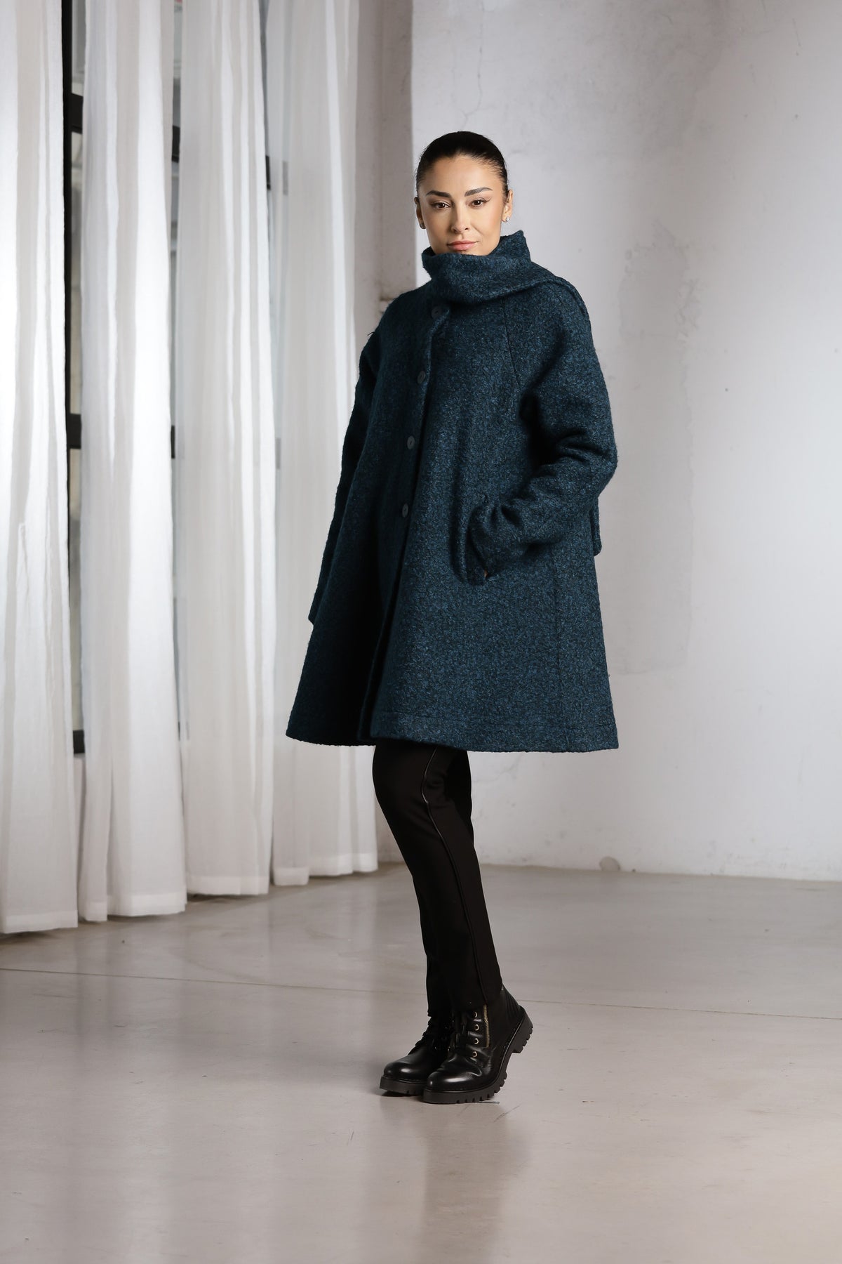 A woman modeling a dark teal textured coat with a high mock neck, paired with black tights and black leather shoes, standing in a minimalist studio with white curtains.
