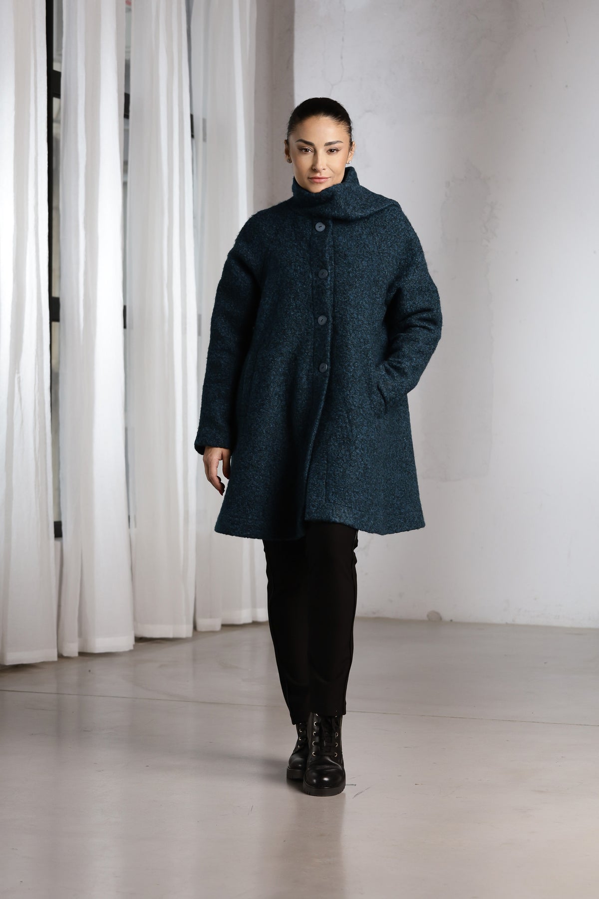 A woman modeling a dark teal textured coat with a high collar and front buttons, paired with black pants and boots, standing in a minimalist studio with white curtains and a concrete floor.