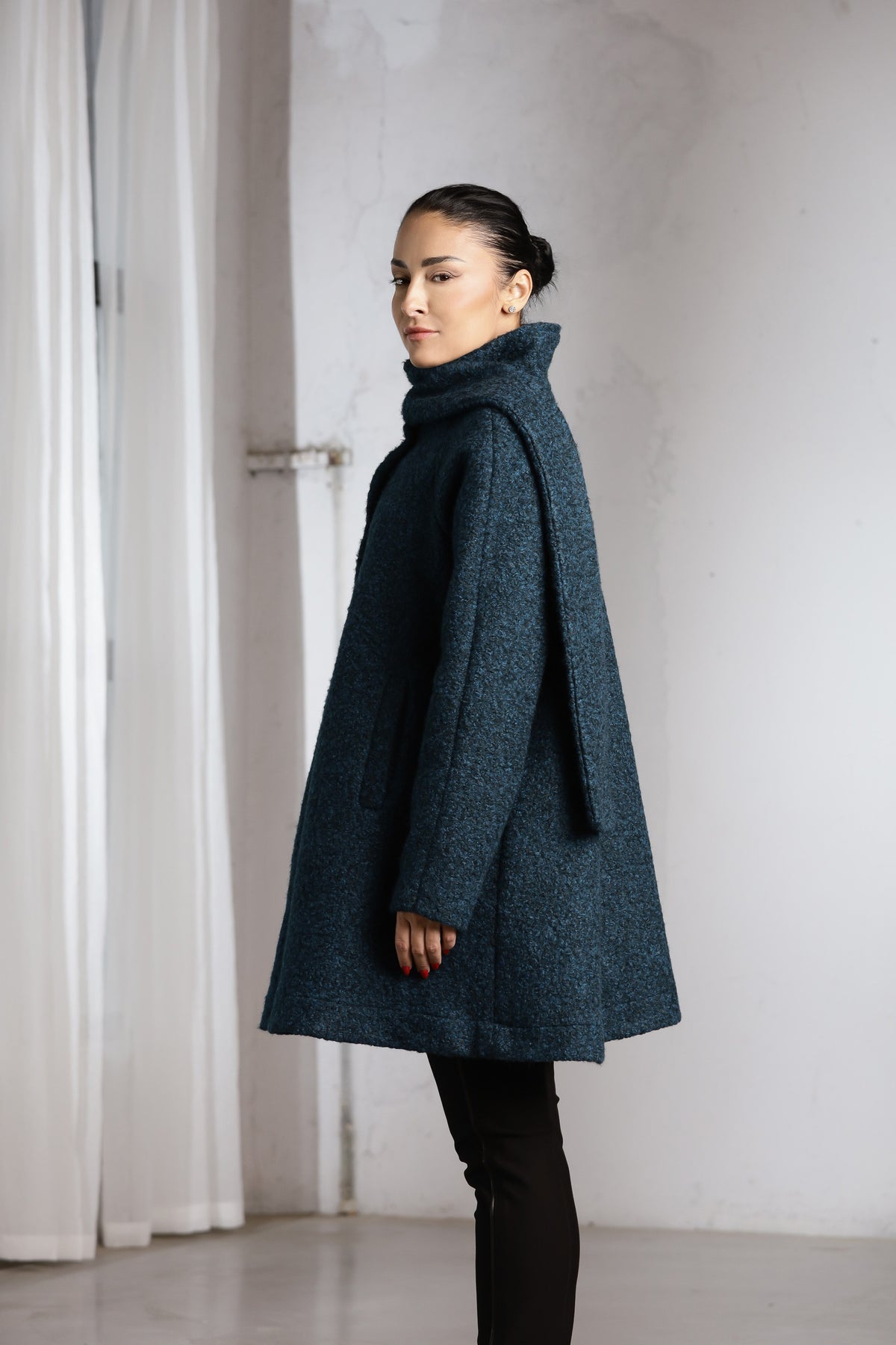 A woman wearing a dark teal textured wool coat with a high folded collar, paired with black pants, standing in a minimalist white room with a curtain on the left.