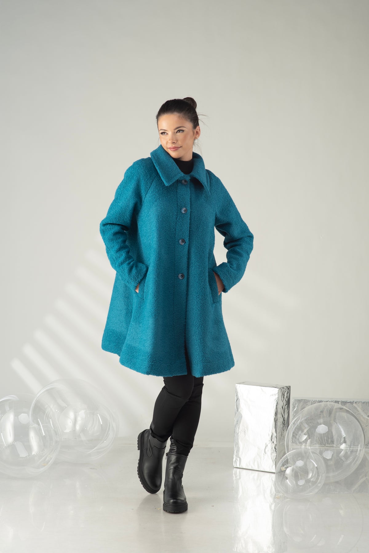 A woman modeling a teal blue wool-blend coat with a high collar and front buttons, paired with black tights and black boots, standing in a studio setting with decorative clear plastic spheres and a silver box.