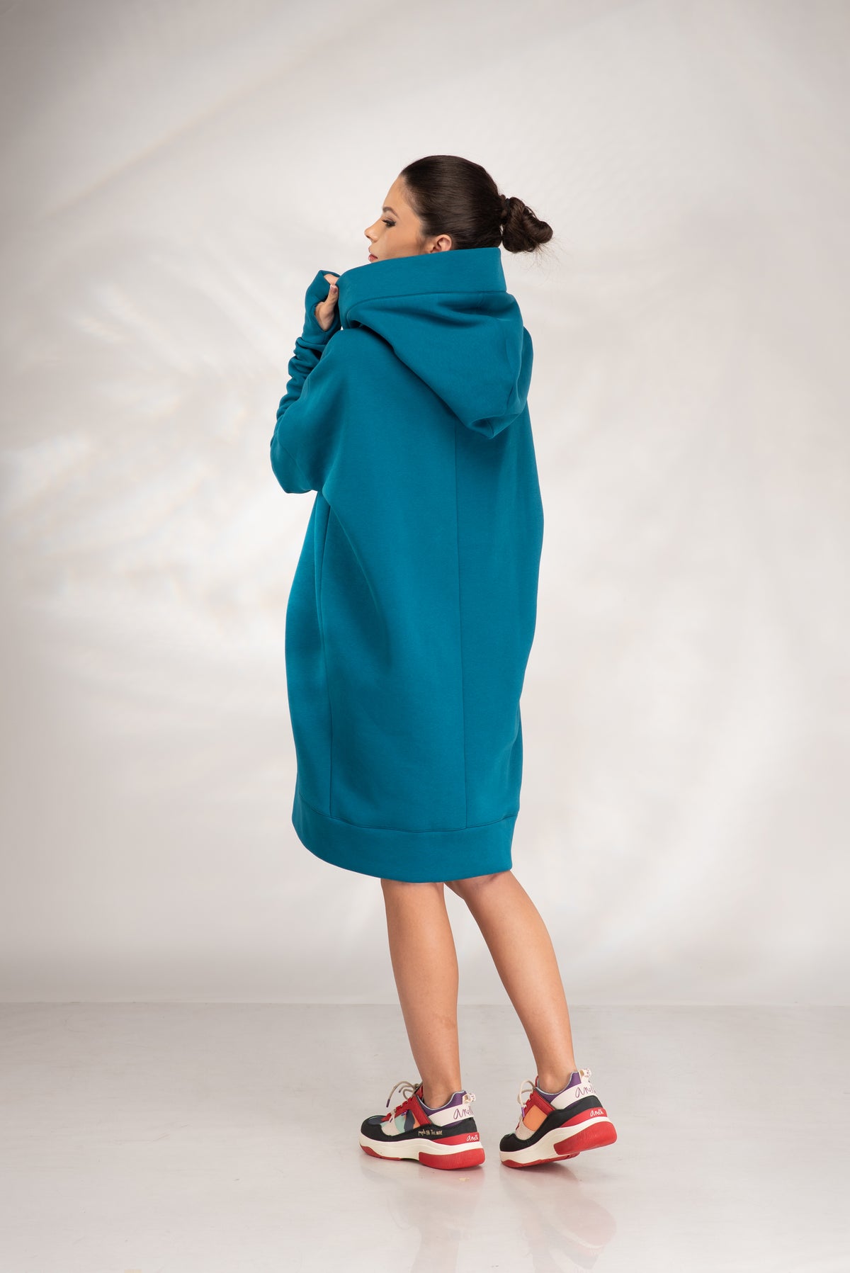 A woman wearing a teal oversized hoodie dress, shown from the back and side against a light gray studio backdrop.
