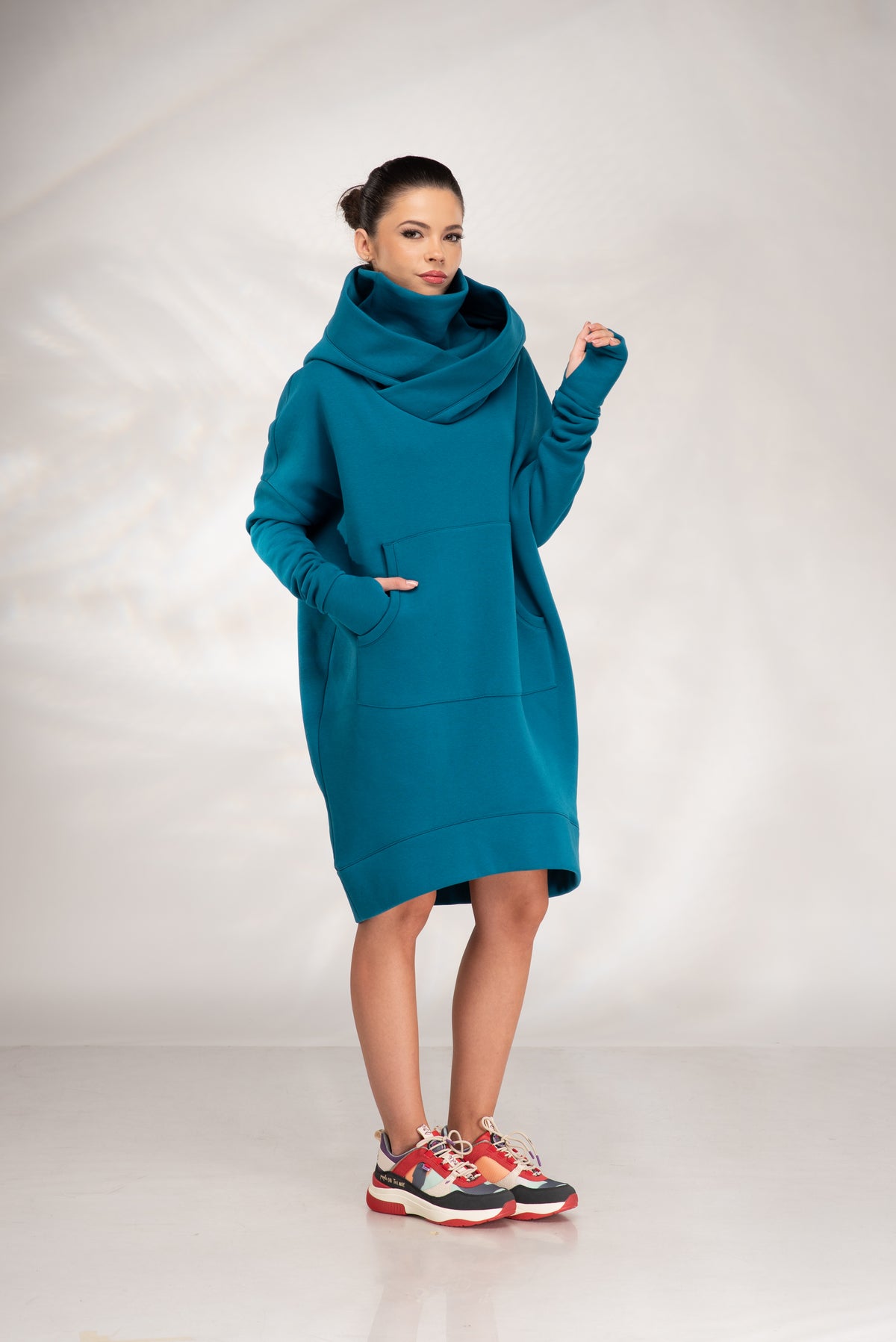 A woman modeling an oversized teal blue-green cowl-neck pullover dress with front pockets, paired with colorful sneakers, against a light gray studio backdrop.