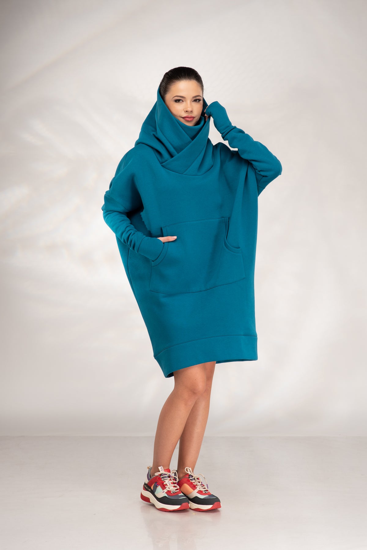 A woman modeling a teal oversized cowl-neck hoodie dress with side pockets, paired with red and gray sneakers, against a light gray studio backdrop.