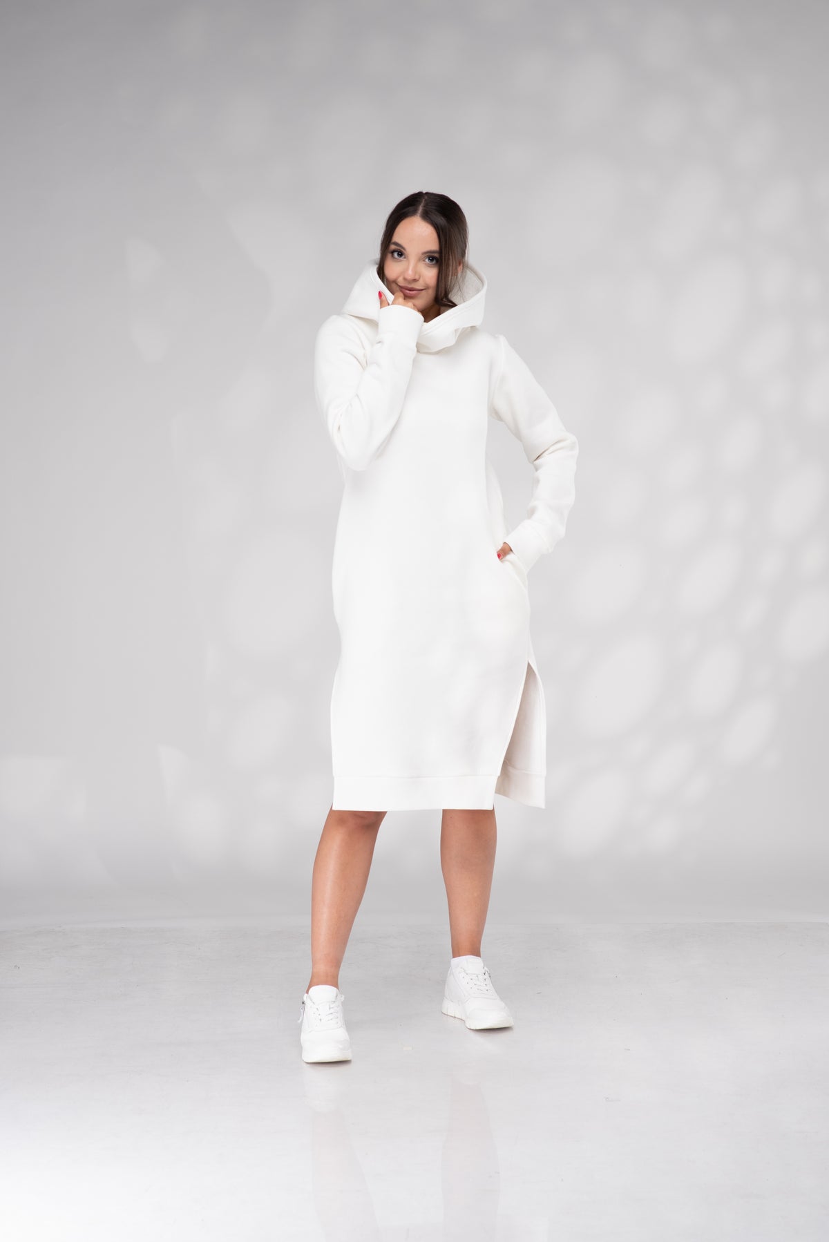 A woman modeling a long white hooded dress with a high cowl neck and side slit, paired with white sneakers, against a light gray studio backdrop with soft circular light patterns.
