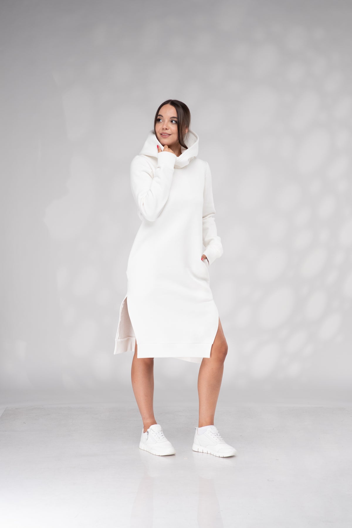 A woman modeling a white hooded knit midi dress with side slits, paired with white sneakers, standing against a light gray studio backdrop with soft shadow patterns.