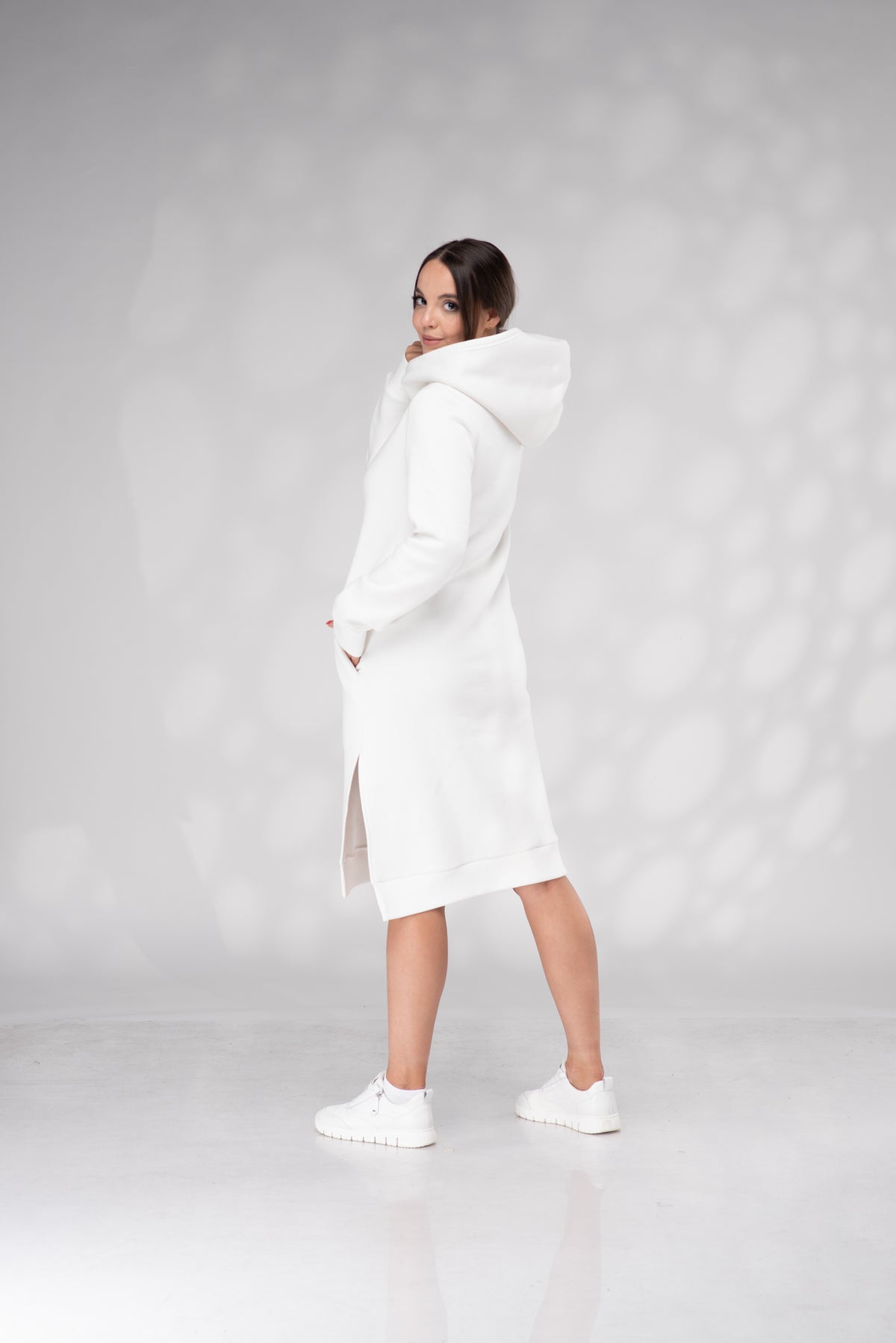 A woman modeling a long white hooded coat or dress with side slits, paired with white sneakers, standing against a light gray studio backdrop with soft circular light patterns.