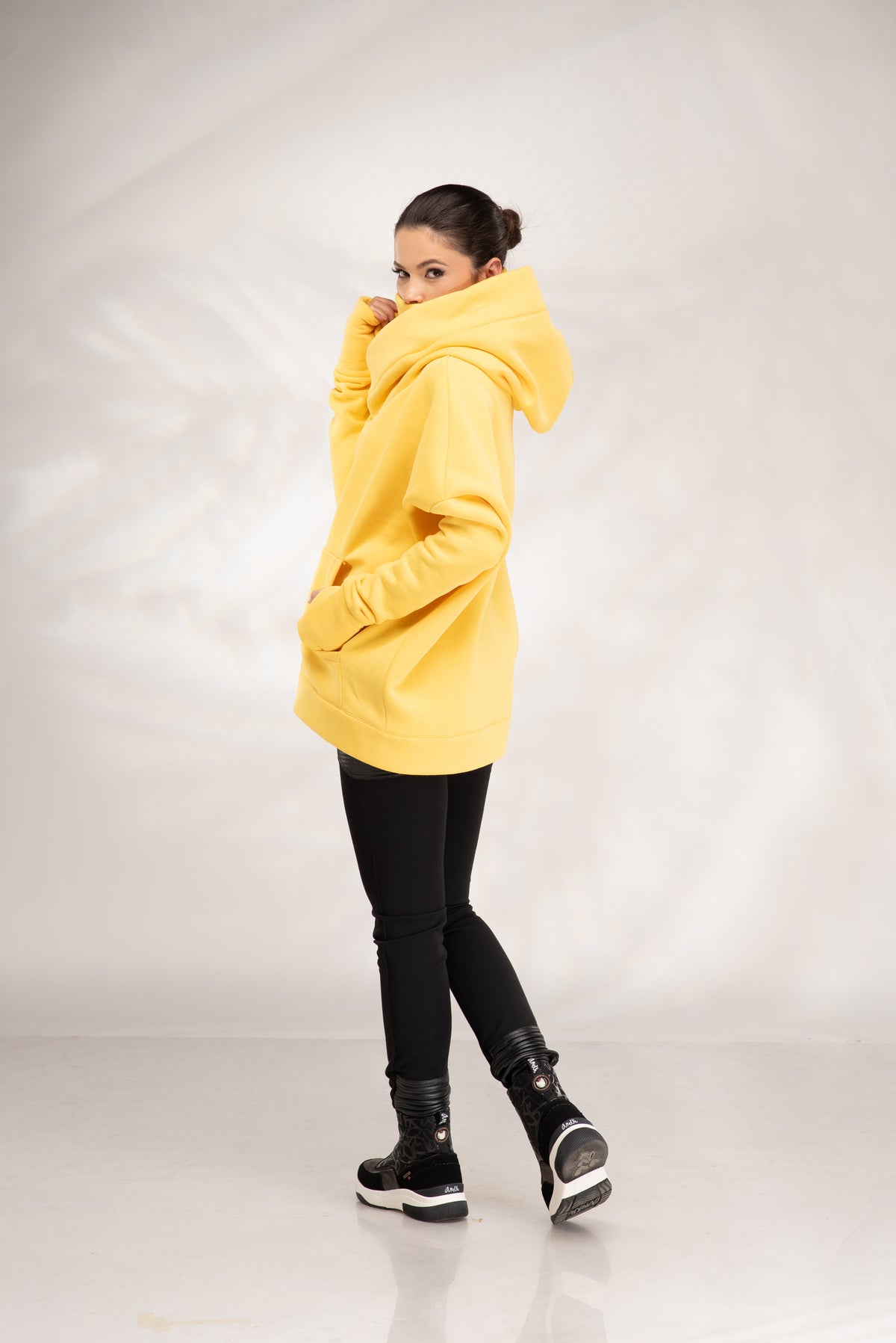 A woman modeling a bright yellow oversized hoodie, paired with black skinny pants and black-and-white sneakers, standing against a light gray studio backdrop.